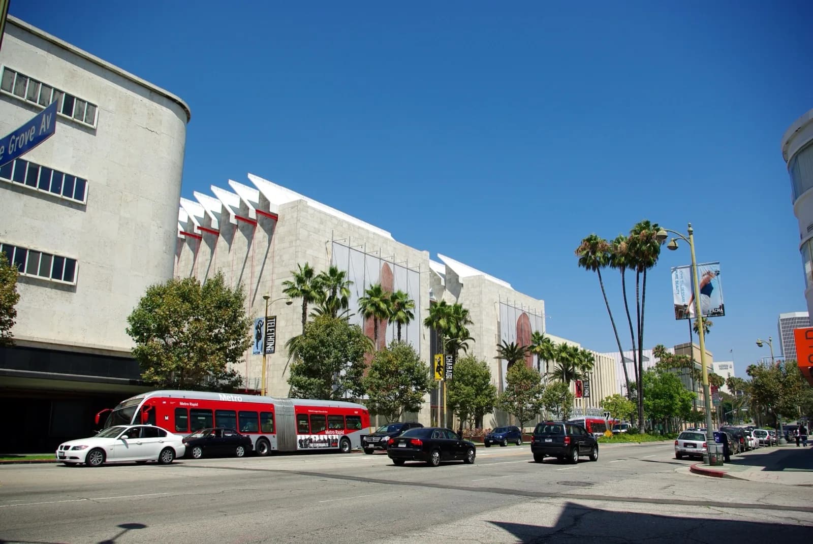 Wide view across Wilshire Boulevard of the Los Angeles County Museum of Art campus, featuring palm trees, museum pavilions with distinctive sawtooth rooflines, and prominent signage under a clear blue sky.