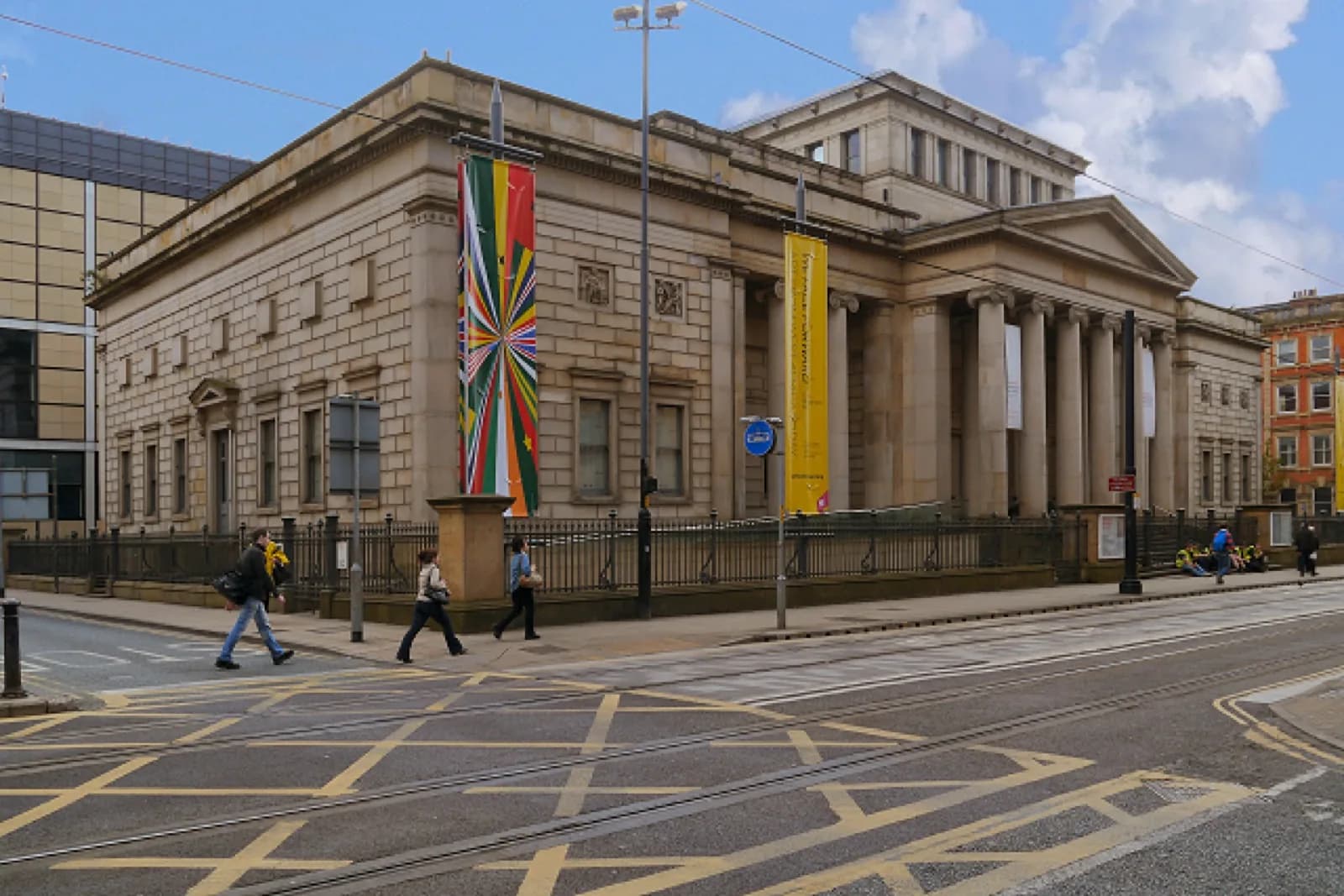 The Manchester Art Gallery building on Mosley Street in Manchester, UK, a neoclassical stone building with columns and pediment, displaying colorful banners on a sunny day.