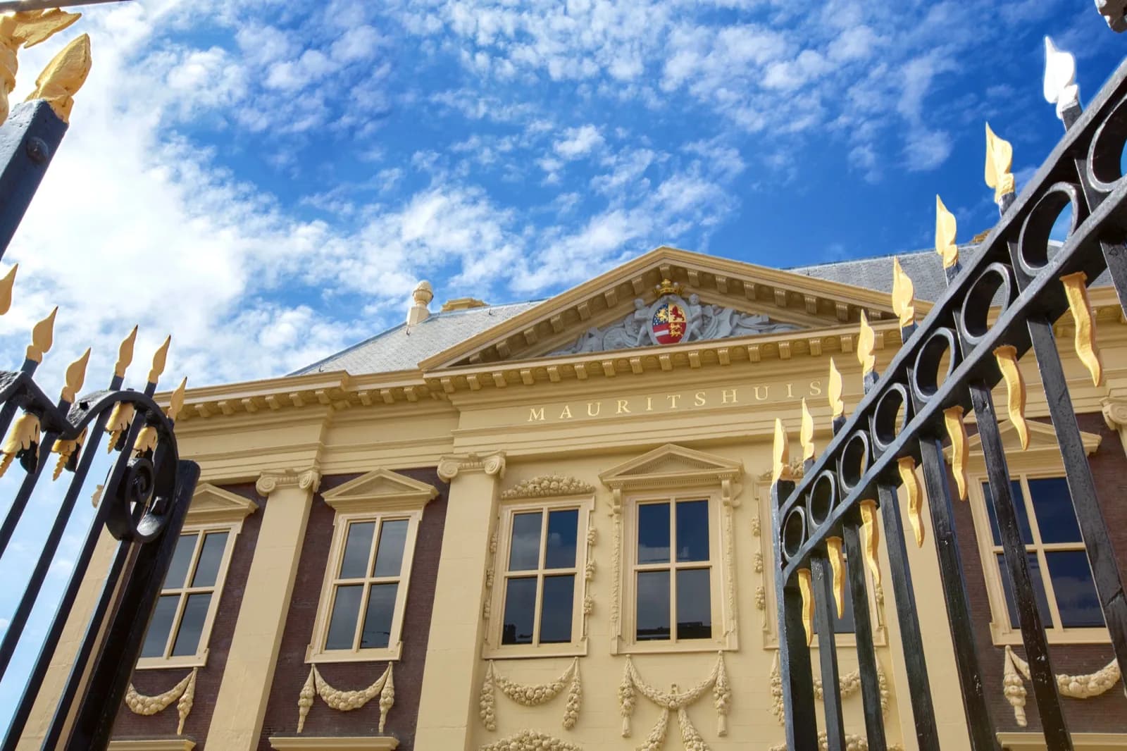 The golden-yellow classical facade of the Mauritshuis museum in The Hague, featuring ornate windows, decorative garlands, a central pediment with coat of arms, and a black wrought-iron fence with gold accents in the foreground under a bright blue sky.