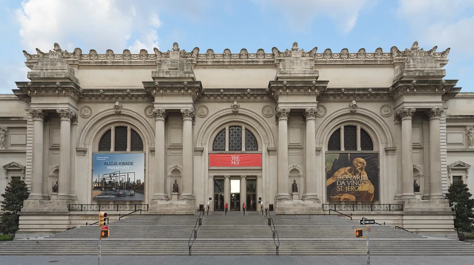 The grand neoclassical facade of The Metropolitan Museum of Art (The Met) in New York City, featuring tall columns, ornate pediment, and wide stone steps leading to the entrance, with exhibition banners under a bright blue sky.