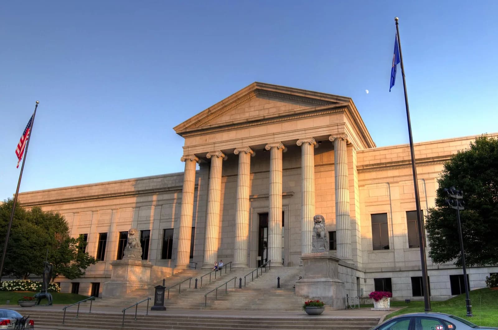 Grand Beaux-Arts facade of the Minneapolis Institute of Art, featuring tall columns, pediment, steps with lion statues, flags, and surrounding trees under a clear blue sky at sunset.