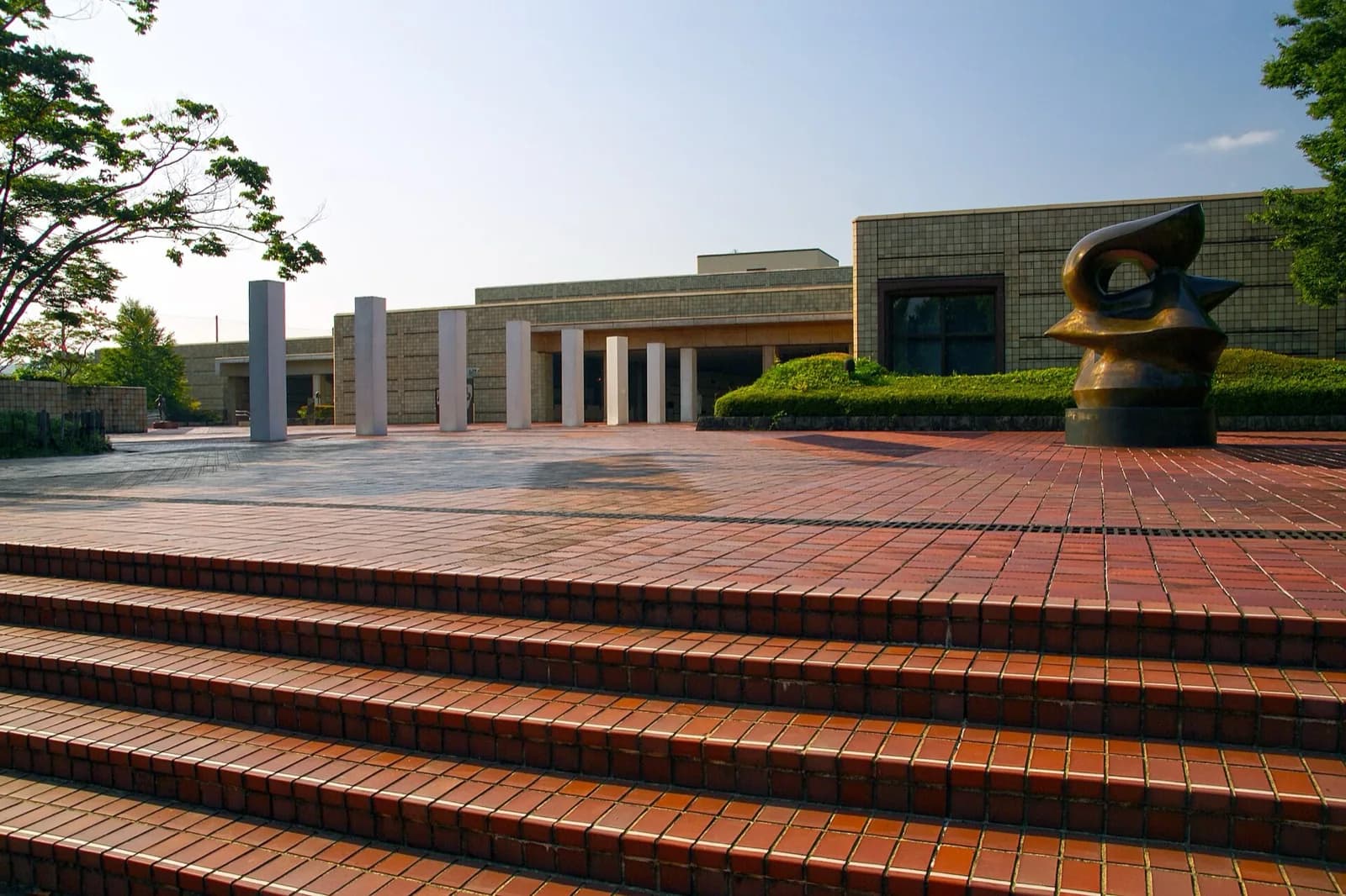 The modern geometric architecture of the Miyagi Museum of Art in Sendai, Japan, with its white and gray forms, large cantilevered roof, and surrounding trees and sculpture garden under a bright blue sky.