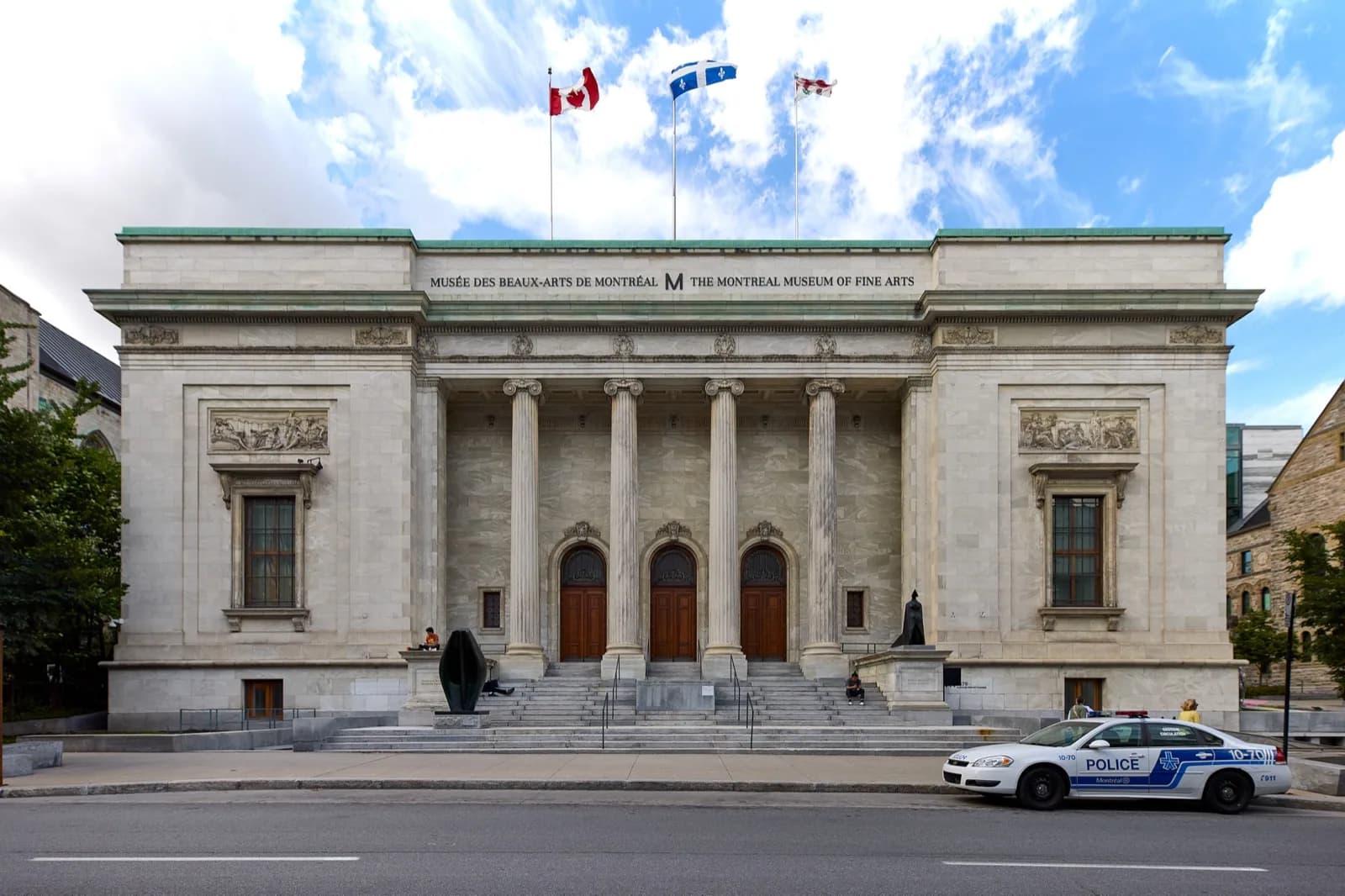 Neoclassical stone facade of the Michal and Renata Hornstein Pavilion at the Montreal Museum of Fine Arts on Sherbrooke Street West, featuring tall fluted columns, pediment with bilingual signage, arched doorways, sculptures, steps, and Canadian and Quebec flags flying against a blue sky.