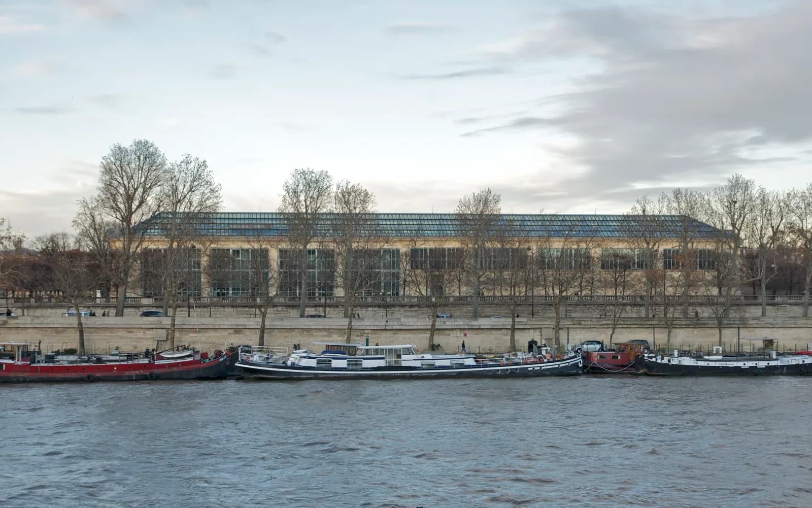 The Musée de l'Orangerie in the Tuileries Garden in Paris, a long neoclassical orangery building with glass roof, viewed across the Seine River with bare trees and houseboats in the foreground under an overcast sky.