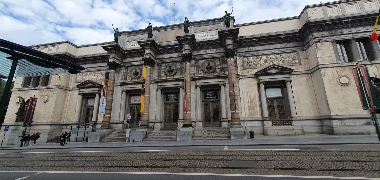 The grand ornate stone facade of the Musées Royaux des Beaux-Arts in Brussels, featuring classical columns, pediments, sculptural reliefs and rooftop statues under a partly cloudy sky, with tram tracks in the foreground.