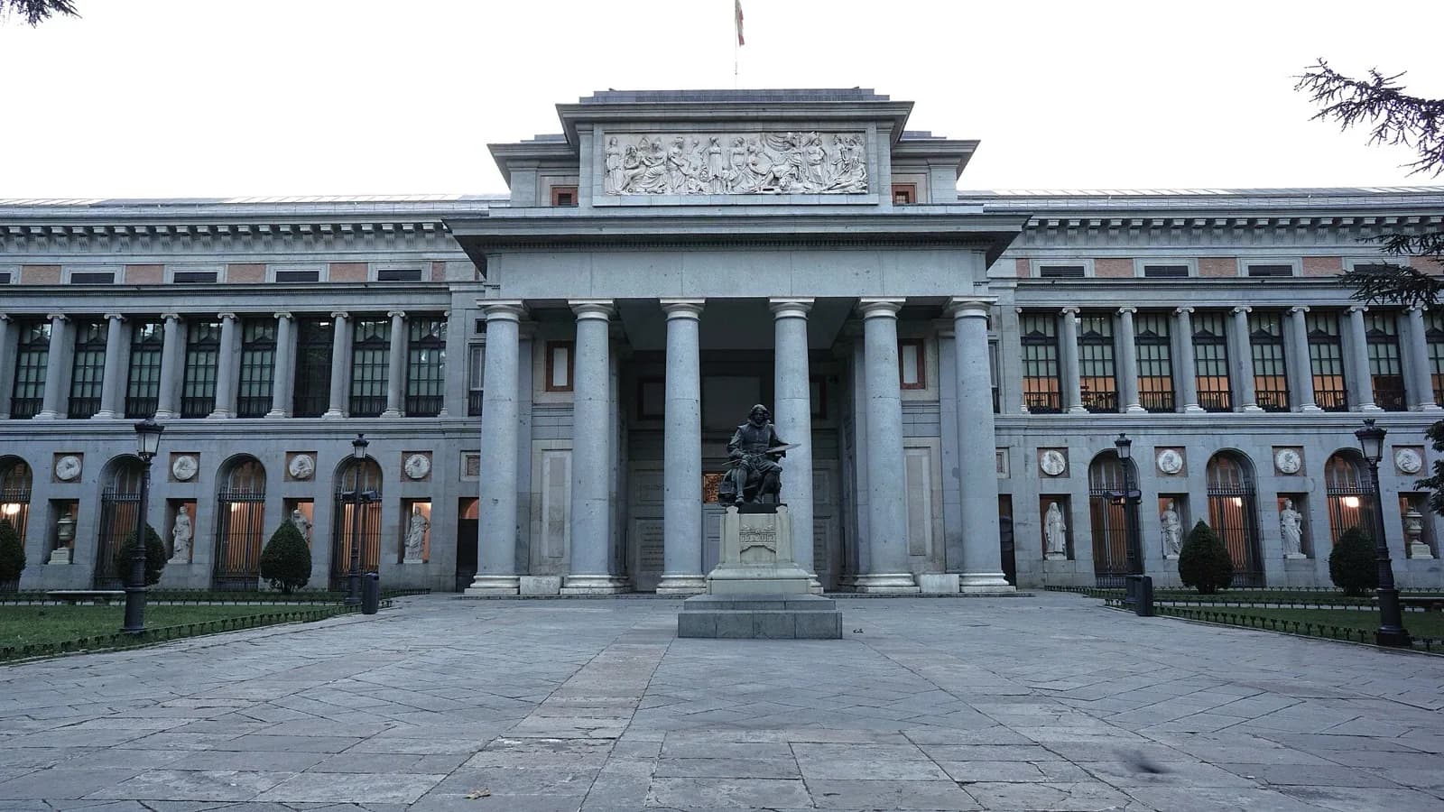 The main neoclassical façade of the Museo Nacional del Prado in Madrid, Spain, designed by Juan de Villanueva, featuring a grand colonnade, pediment with sculptures, central fountain and surrounding trees under a blue sky.