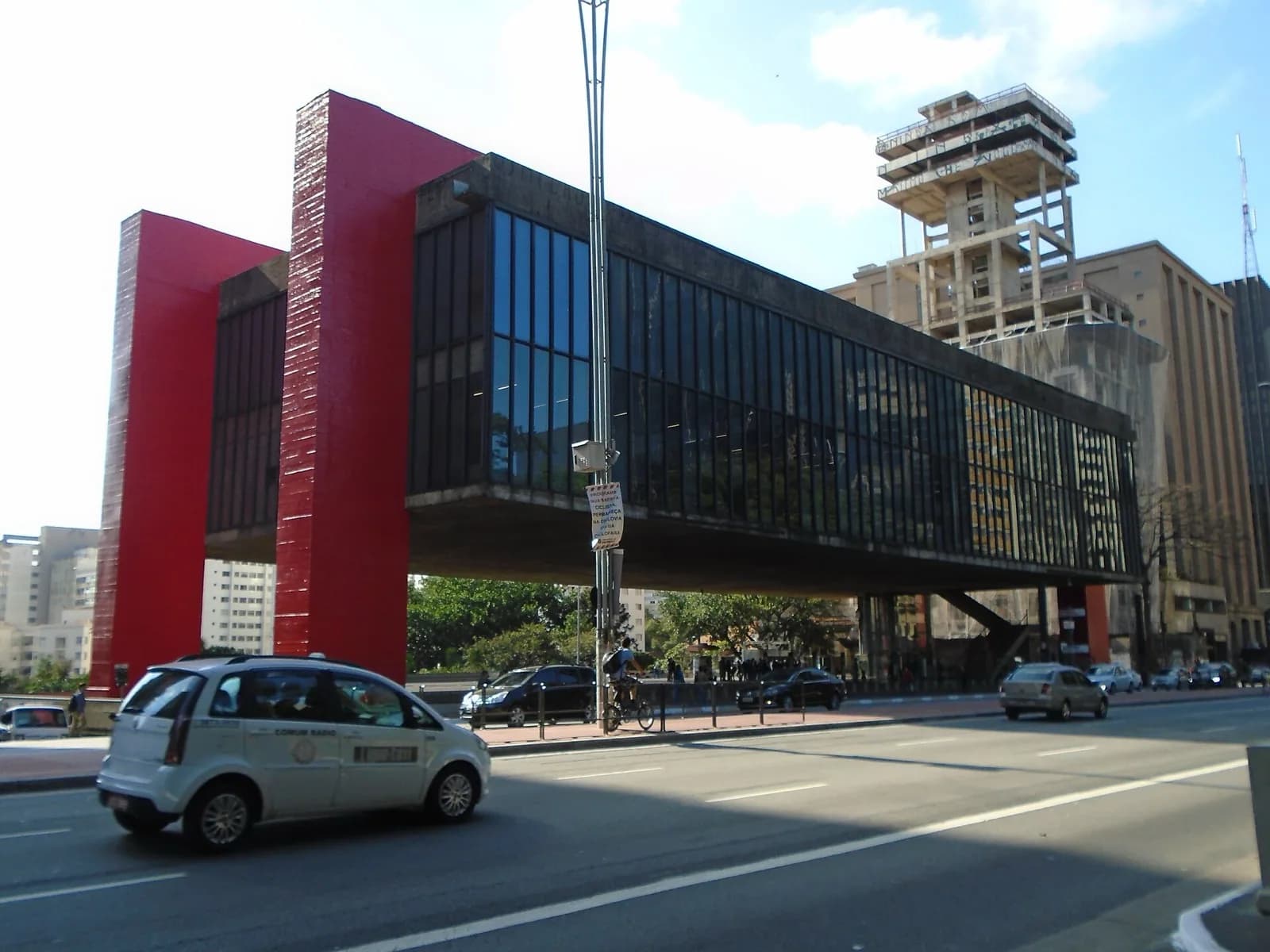 The iconic suspended concrete structure of the Museu de Arte de São Paulo (MASP) on Avenida Paulista, a brutalist building by Lina Bo Bardi elevated on two massive red pillars, with traffic and city life below under a bright sky.