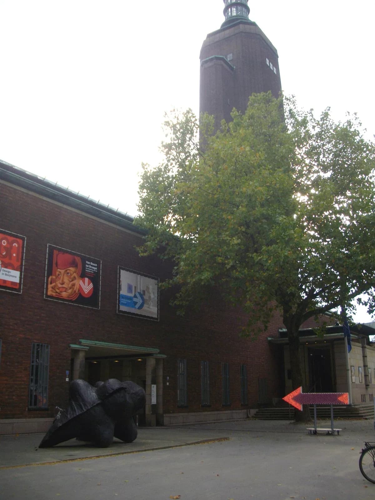 The brick facade and main entrance of Museum Boijmans Van Beuningen in Rotterdam, Netherlands, featuring large colorful exhibition banners including one for "Van Bosch tot Bruegel", a prominent abstract black sculpture in the foreground, entrance columns, trees, and a tall brick tower in the background under an overcast sky.
