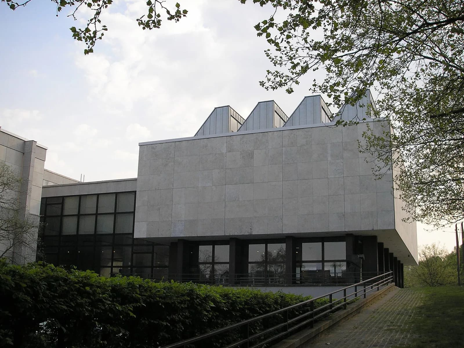 The modern low-rise buildings of Museum Dahlem (part of the Staatliche Museen zu Berlin) with large windows and flat roofs, set amid trees and lawn under an overcast sky.