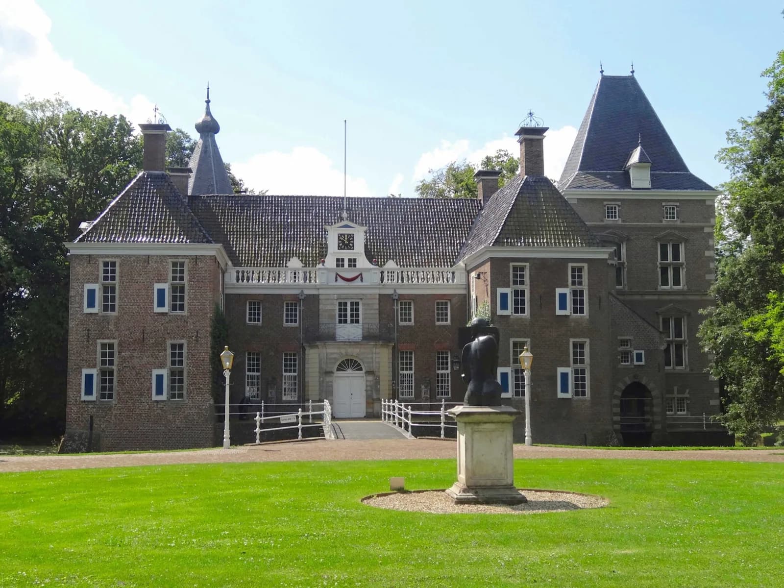 The historic brick facade of Kasteel het Nijenhuis (Museum de Fundatie) near Heino, with twin towers, central clock gable, white drawbridge entrance, and a sculpture on the lawn under a blue sky with clouds, Overijssel, Netherlands.