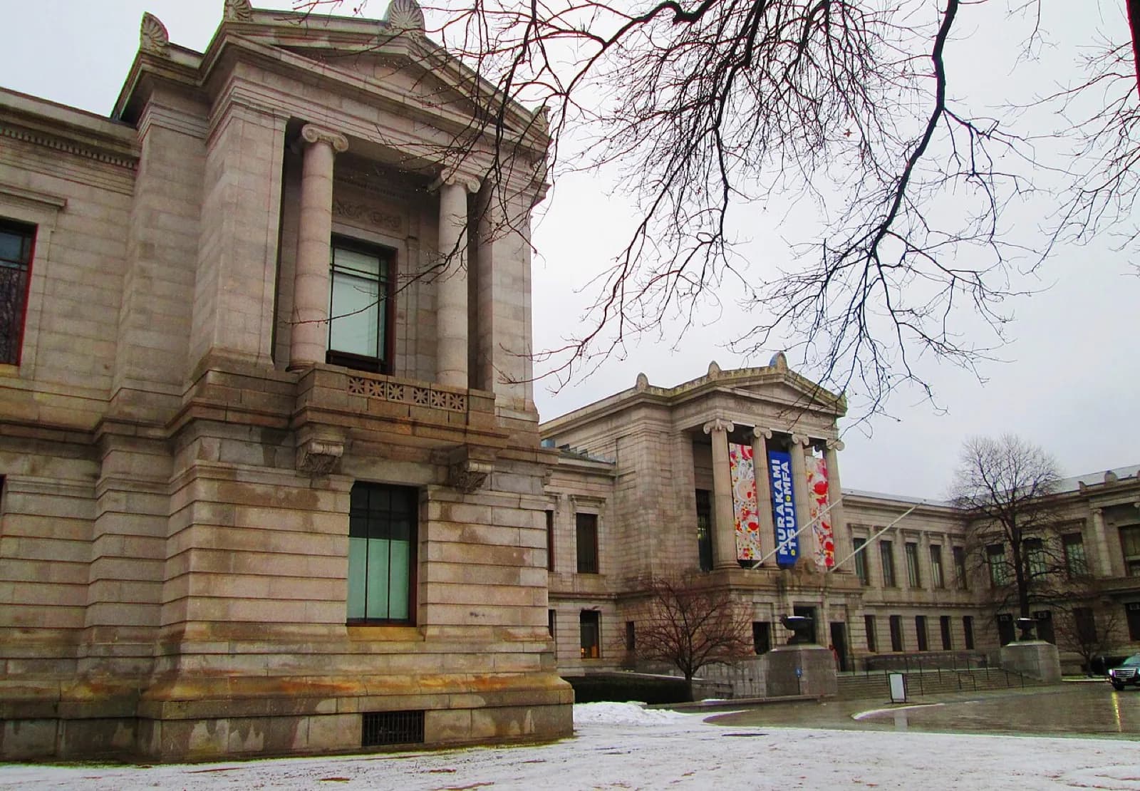 The grand neoclassical stone facade of the Museum of Fine Arts, Boston on Huntington Avenue, featuring tall columns, a pediment, and colorful Yayoi Kusama exhibition banners on the central pavilion, with snow on the ground and bare tree branches in the foreground on an overcast winter day.