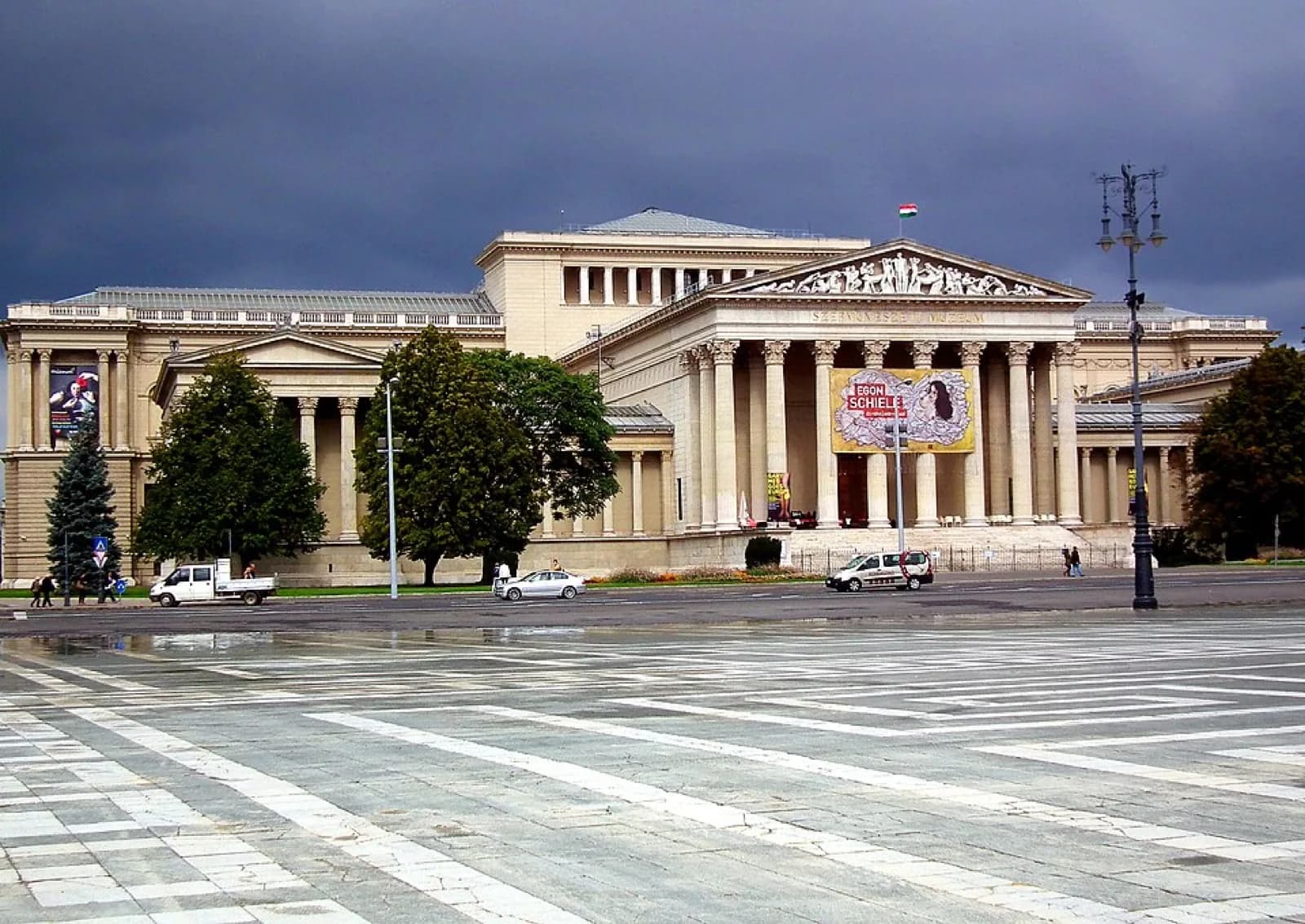 The monumental neoclassical facade of the Museum of Fine Arts (Szépművészeti Múzeum) on Heroes' Square in Budapest, featuring a grand portico with tall columns, pediment sculptures, wide stone steps, and visitors in front of the building under a blue sky.