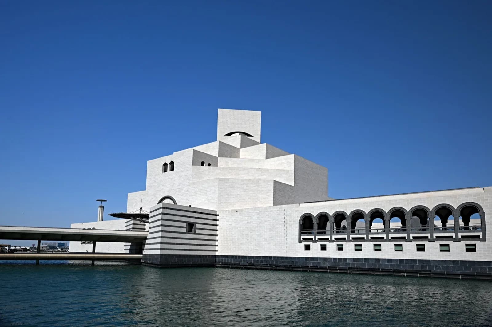 The iconic white limestone geometric building of the Museum of Islamic Art in Doha, Qatar, designed by I. M. Pei, featuring arched loggias, stepped forms, and a bridge, viewed across the water under a deep blue sky.