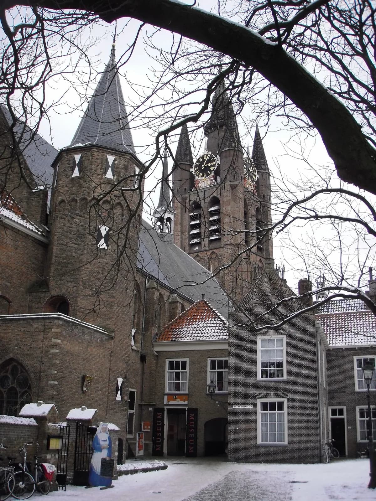 The historic brick facade of Museum Prinsenhof Delft in the Netherlands, a former convent with stepped gables, viewed on a snowy winter day with the tall Gothic towers and clock of the Oude Kerk rising behind it, bare tree branches in the foreground, and a small statue and bicycles near the entrance.