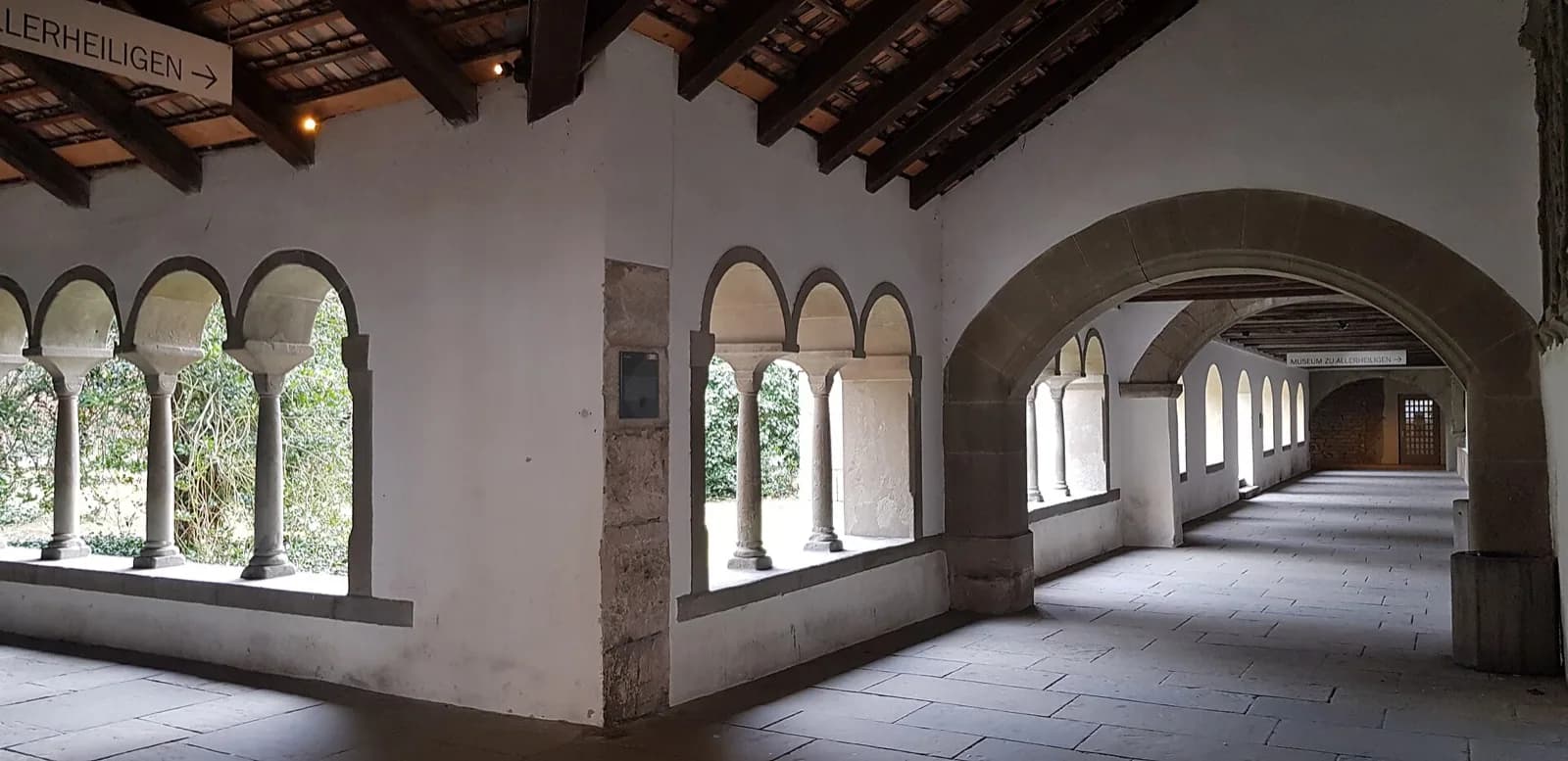 The historic stone cloister arcade with rounded arches at the Museum zu Allerheiligen in Schaffhausen, Switzerland, leading toward the museum entrance under a covered walkway with signs.