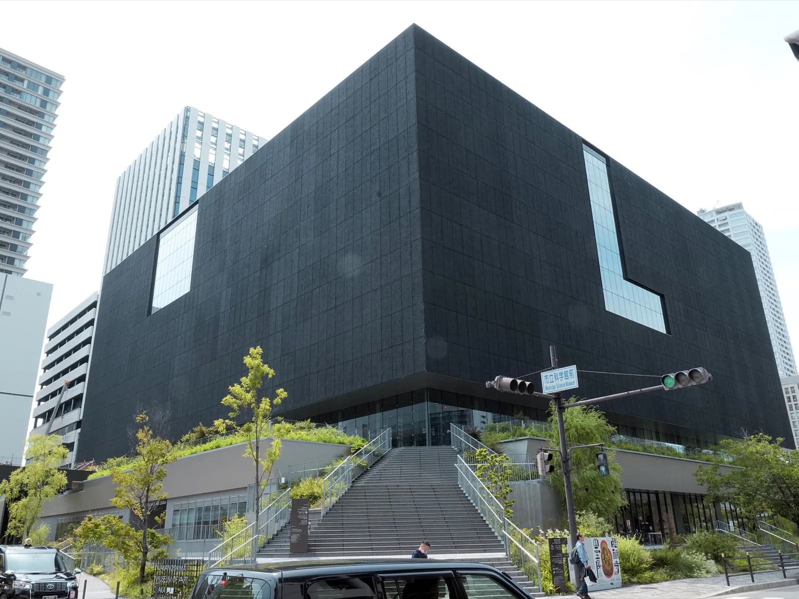 The modern glass-fronted Nakanoshima Museum of Art (NAKKA) in Osaka, Japan, a contemporary building with large windows along the Dōjima River, with a bridge and city skyline visible under a clear blue sky.