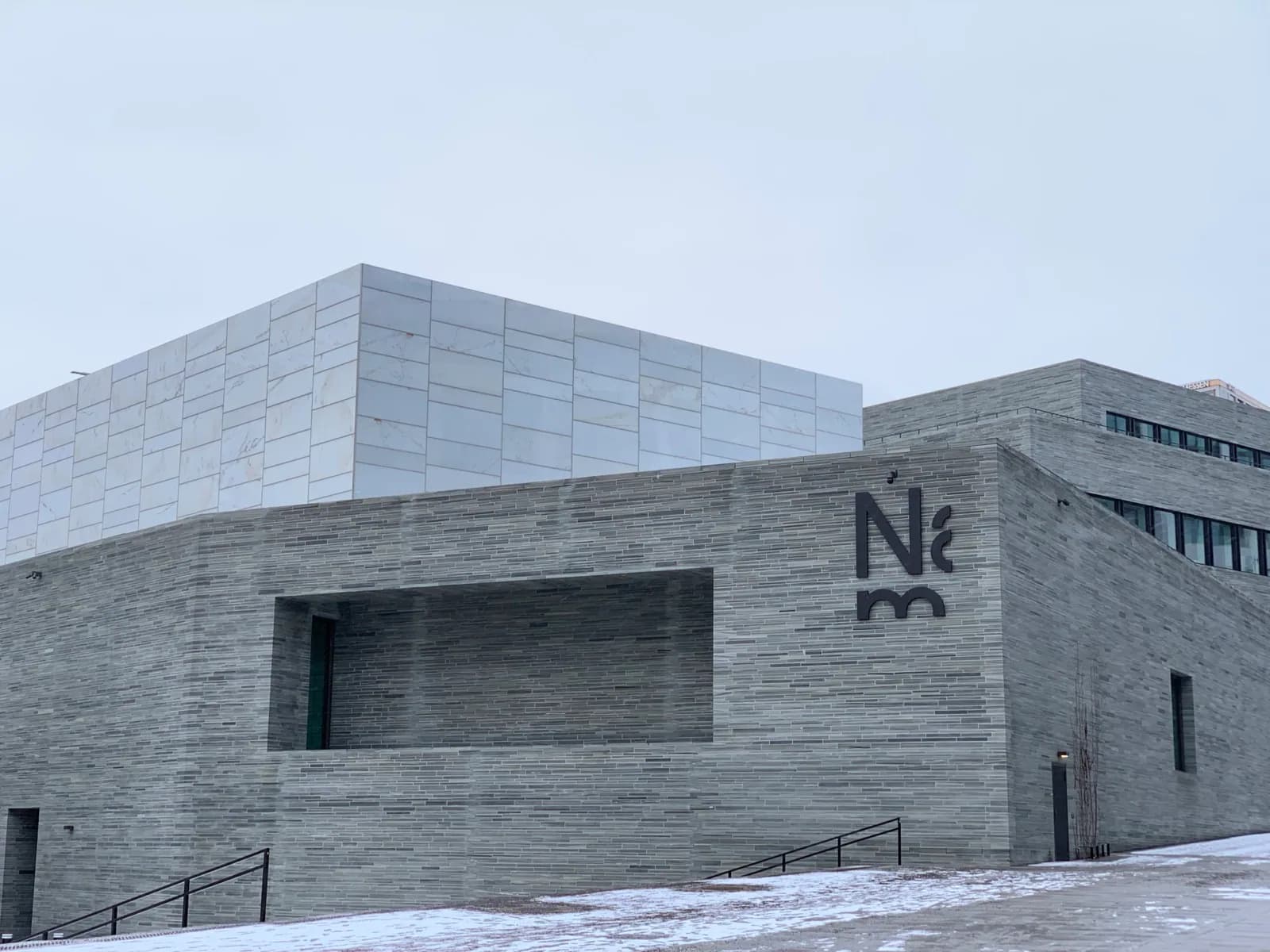 Modern angular facade of the new Nasjonalmuseet in Oslo, clad in gray stone and white marble panels, with large dark 'Nm' logo signage, set against a snowy plaza under an overcast sky.