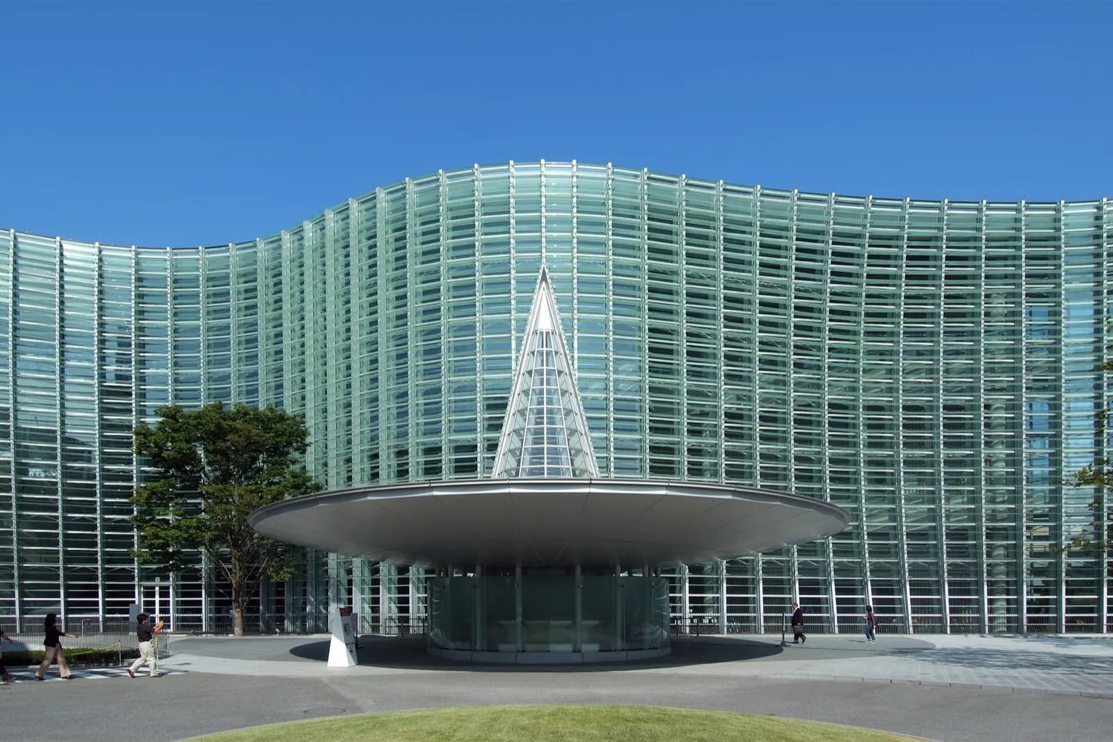 The National Art Center in Tokyo, Japan, a modern building with a large curved glass facade and a distinctive conical glass entrance structure, viewed on a sunny day.