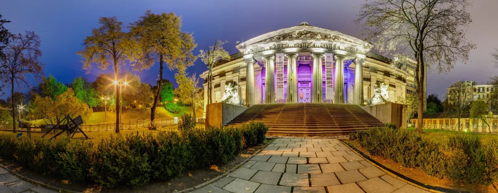 Panoramic view at dusk of the neoclassical National Art Museum of Ukraine in Kyiv, showing its grand columned portico with sculpted pediment, flanked by two large white lion statues on pedestals, wide stone staircase, and surrounding parkland with trees and pathways under a deep blue sky.