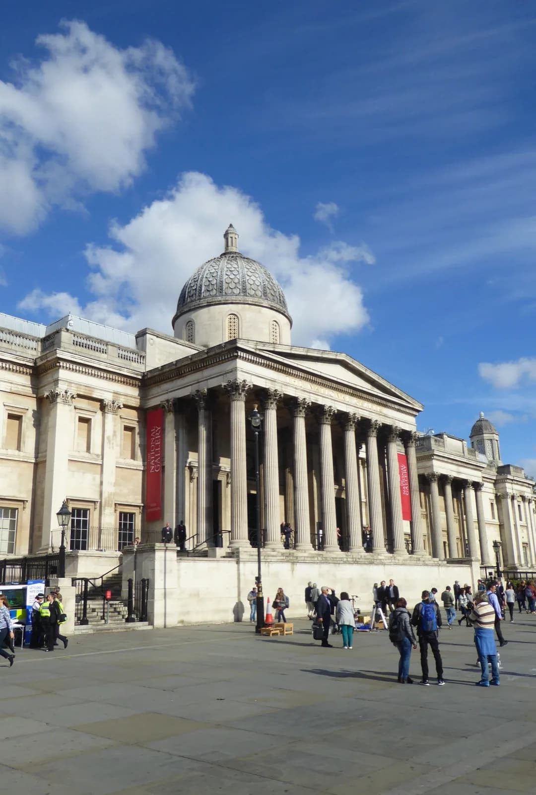 The neoclassical facade of the National Gallery, London, with its grand portico of Corinthian columns supporting a pediment and topped by a prominent dome, viewed from Trafalgar Square on a bright day with visitors in the paved forecourt.