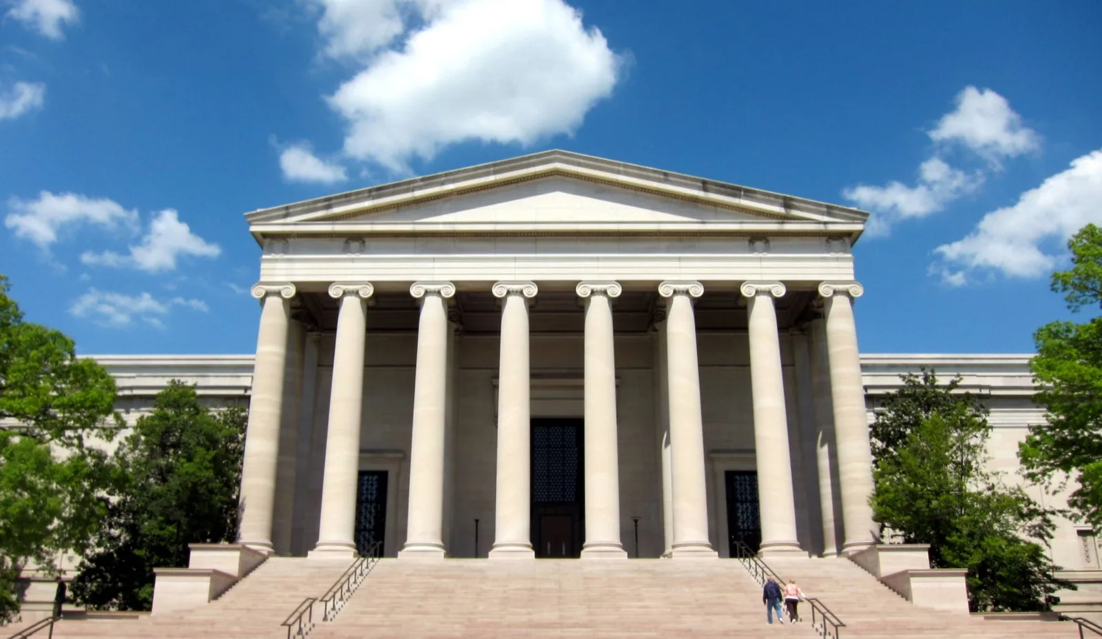 The neoclassical West Building of the National Gallery of Art on the National Mall in Washington, D.C., featuring a grand portico with Ionic columns and wide stone steps under a bright blue sky.