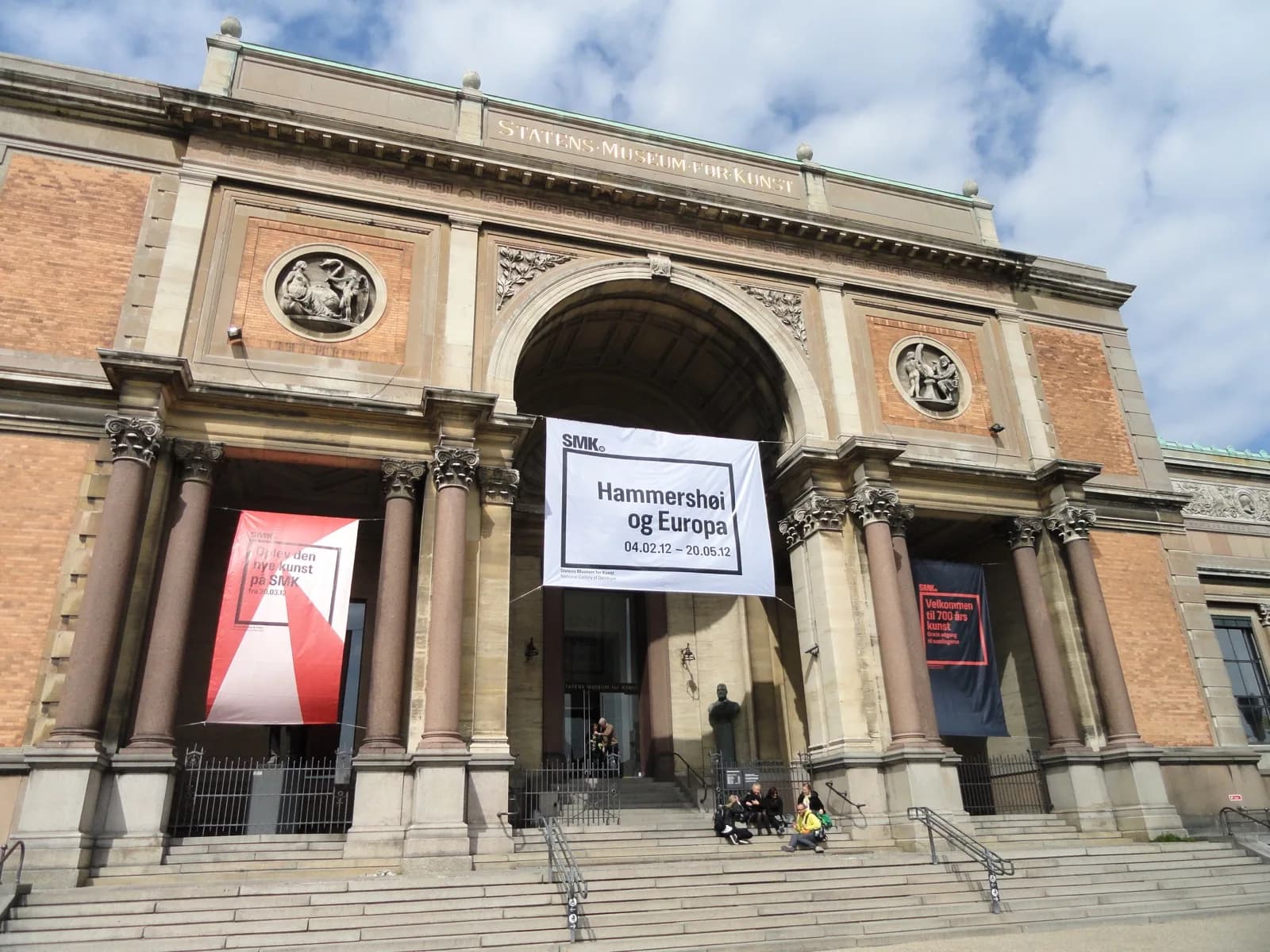 Main entrance facade of Statens Museum for Kunst (National Gallery of Denmark) in Copenhagen, featuring a grand arched portico with columns, brick and stone details, inscribed pediment, exhibition banners, and visitors on the steps under a partly cloudy sky.