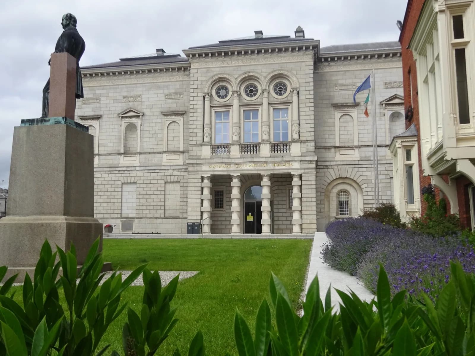 Stone facade of the National Gallery of Ireland on Merrion Square, Dublin, with classical columns, arched windows, pediment details, a foreground statue, and Irish flag under an overcast sky.