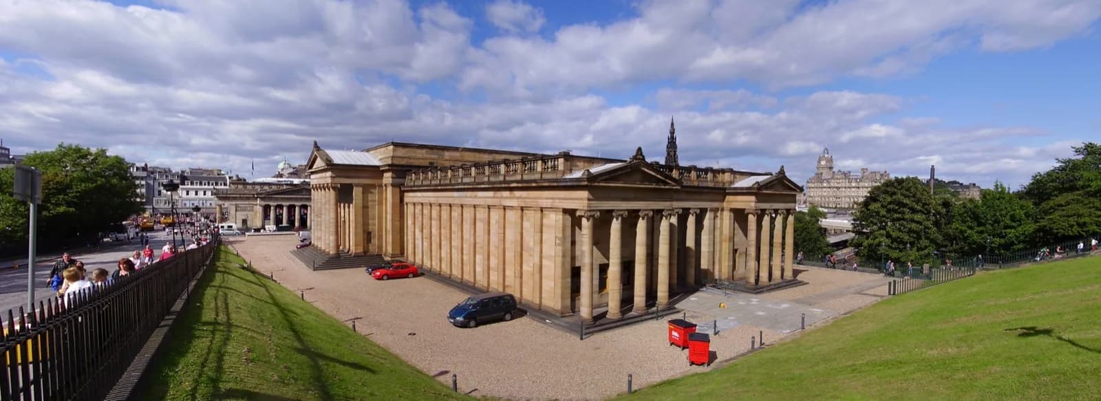 Wide view of the neoclassical National Gallery of Scotland building with its grand columns and pediment on the Mound in Edinburgh, on a sunny day with a blue sky, people and red post boxes in the foreground, and the city skyline with a church spire in the background.