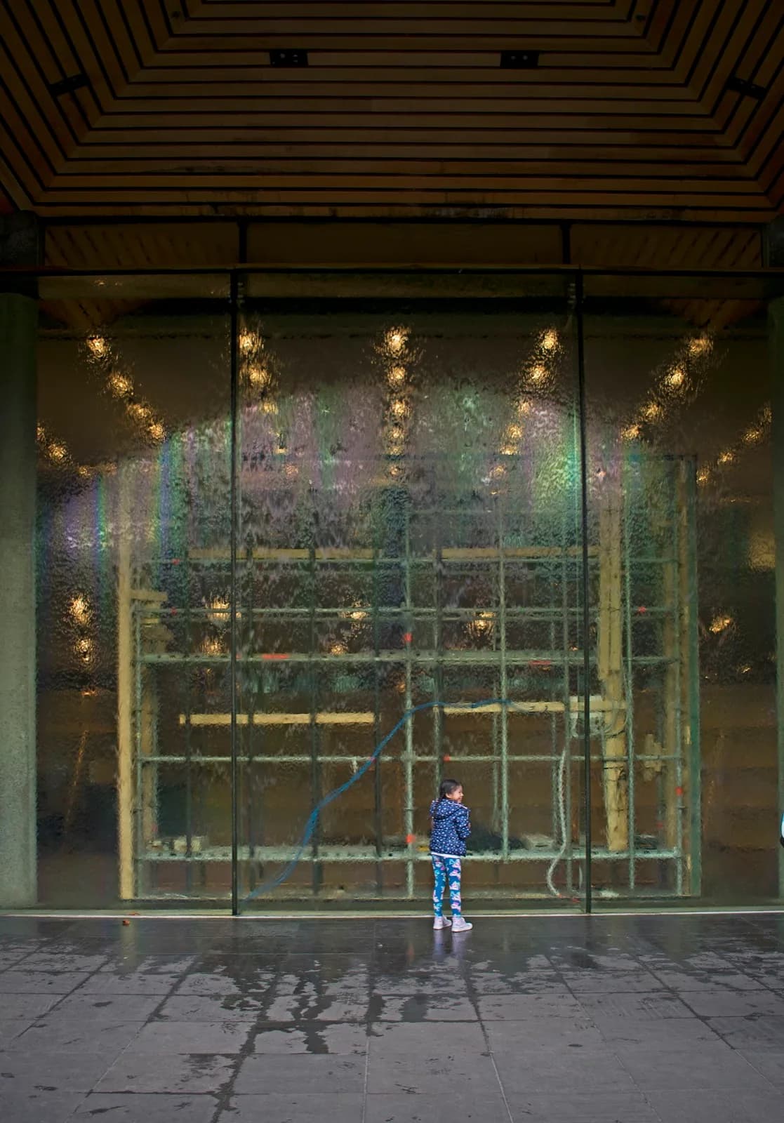 The main façade of the National Gallery of Victoria (NGV) in Melbourne, Australia, featuring a monumental water wall, classical columns, signage and the entrance plaza with trees under a clear blue sky.