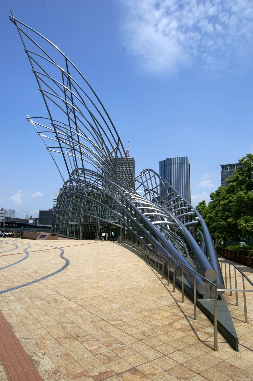 The modern entrance to the National Museum of Art, Osaka (NMAO) on Nakanoshima island, featuring a dramatic steel sculpture and glass pavilion with city skyline in the background under a blue sky.