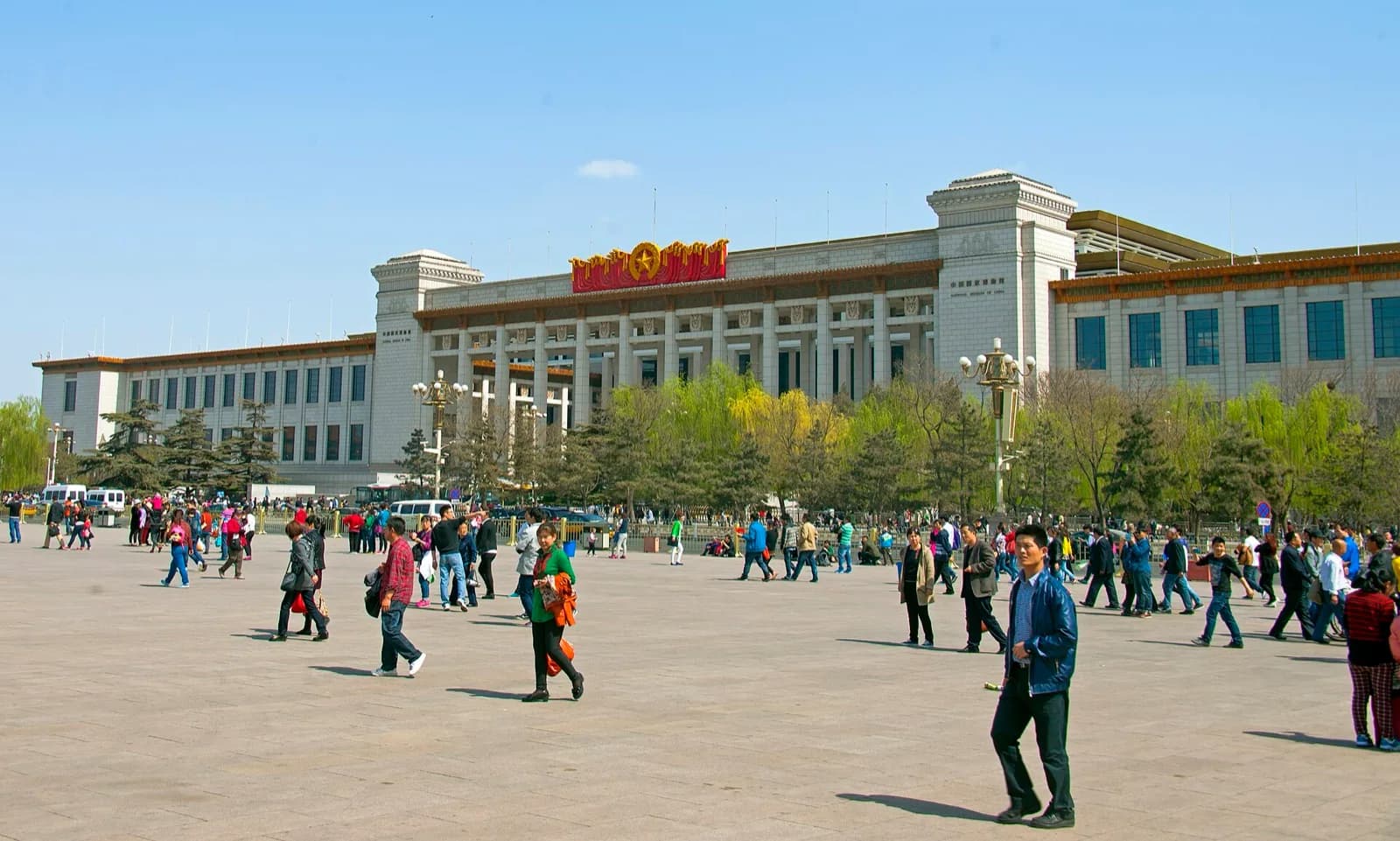 The monumental facade of the National Museum of China on the eastern side of Tiananmen Square in Beijing, a grand classical building with columns and traditional Chinese architectural details under a bright blue sky, with crowds of visitors crossing the expansive paved plaza on a sunny day with spring foliage on the trees.