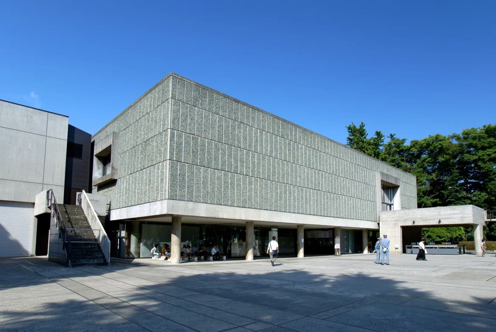 The National Museum of Western Art in Tokyo, a modernist concrete building designed by Le Corbusier with a distinctive green pebble-textured facade, viewed across a sunny plaza with visitors and security guards under a clear blue sky.