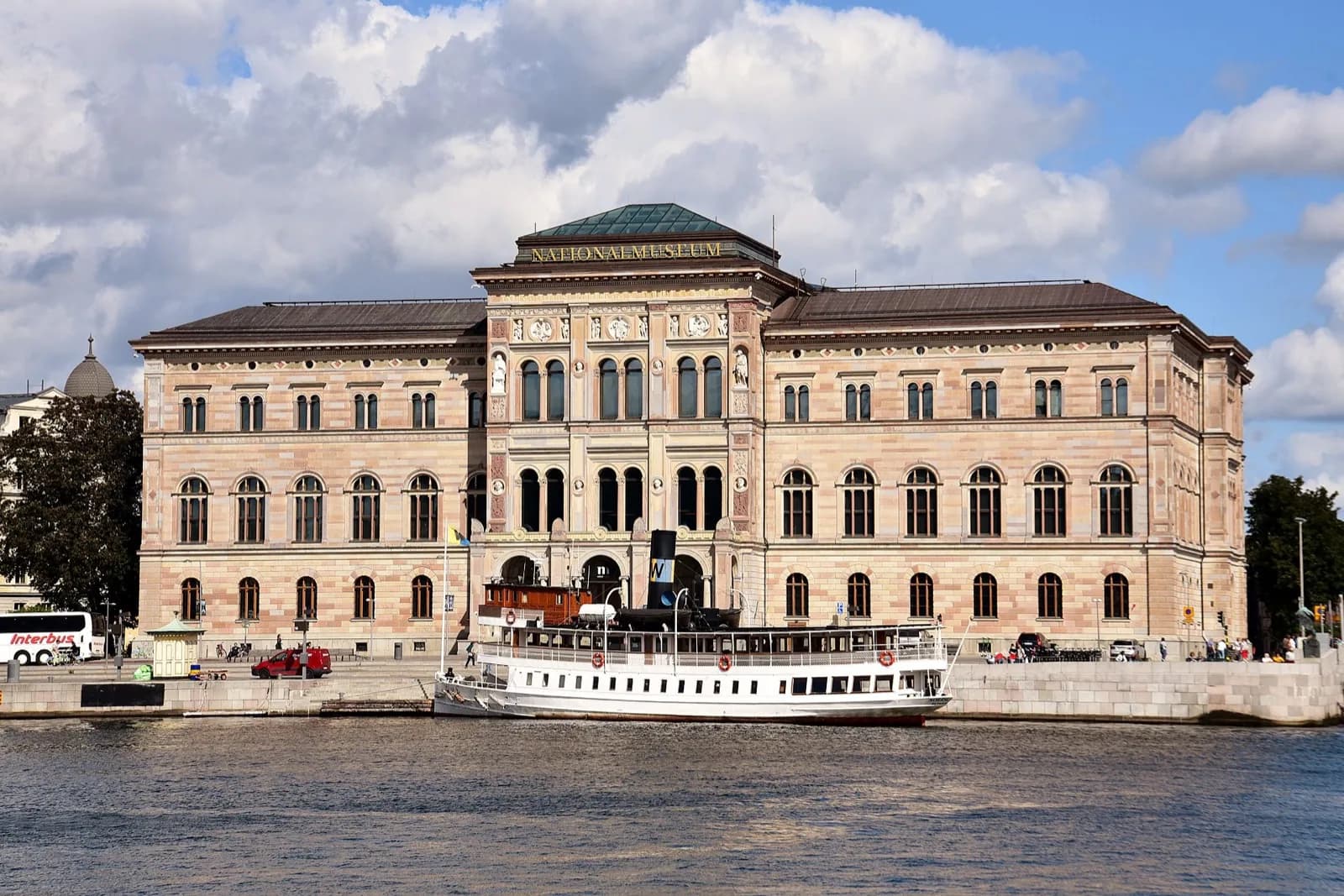 Front facade of the Nationalmuseum, a grand Renaissance Revival stone building with arched windows, columns and sculptures, viewed from across the water with a white steamboat in the foreground, Stockholm, Sweden.