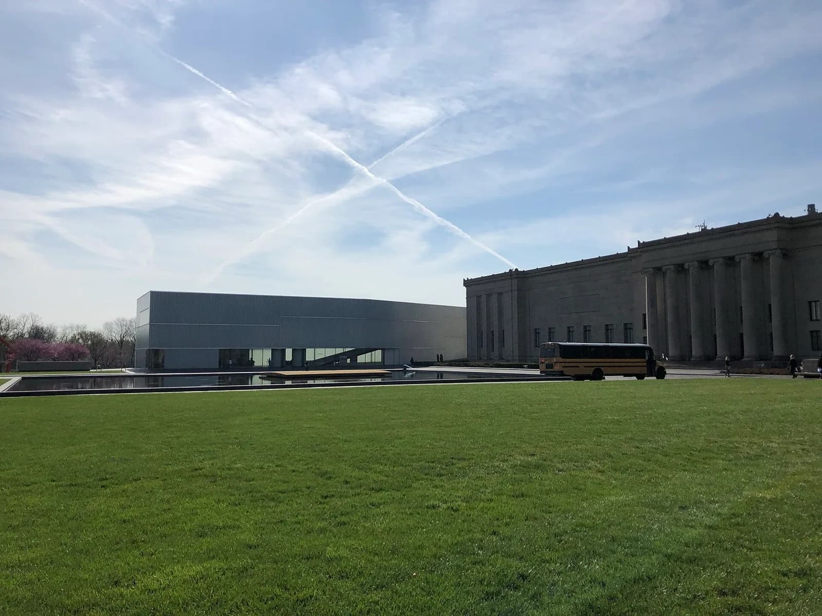 The Nelson-Atkins Museum of Art in Kansas City, showing the original neoclassical building with tall columns beside the modern translucent Bloch Building addition by Steven Holl, reflected in a long pool on a green lawn under a blue sky with contrails, and a school bus parked nearby.