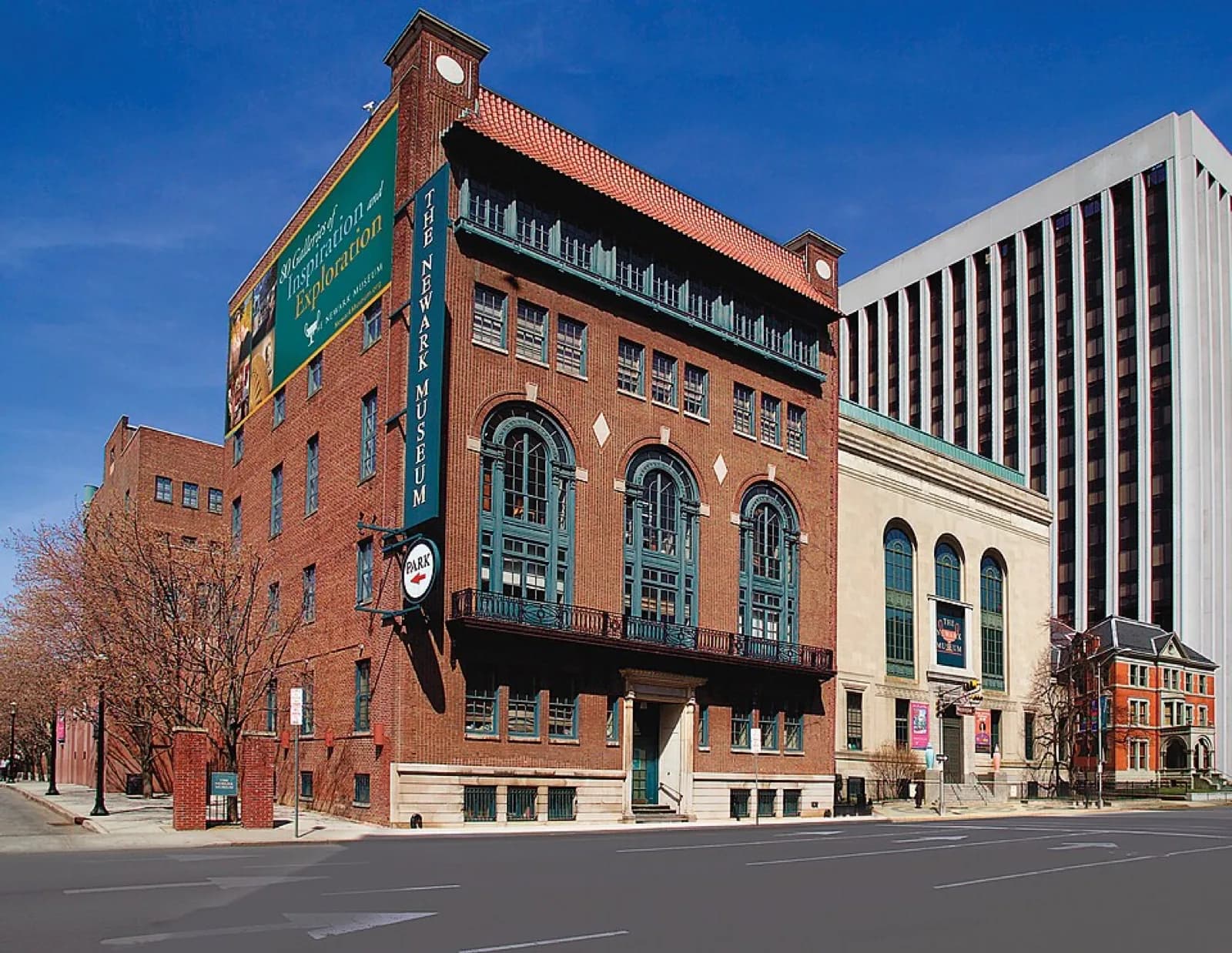The contemporary facade of the Newark Museum of Art in Newark, New Jersey, featuring a glass-fronted entrance wing with the museum name signage, brick elements, and visitors on the sidewalk under a clear blue sky.