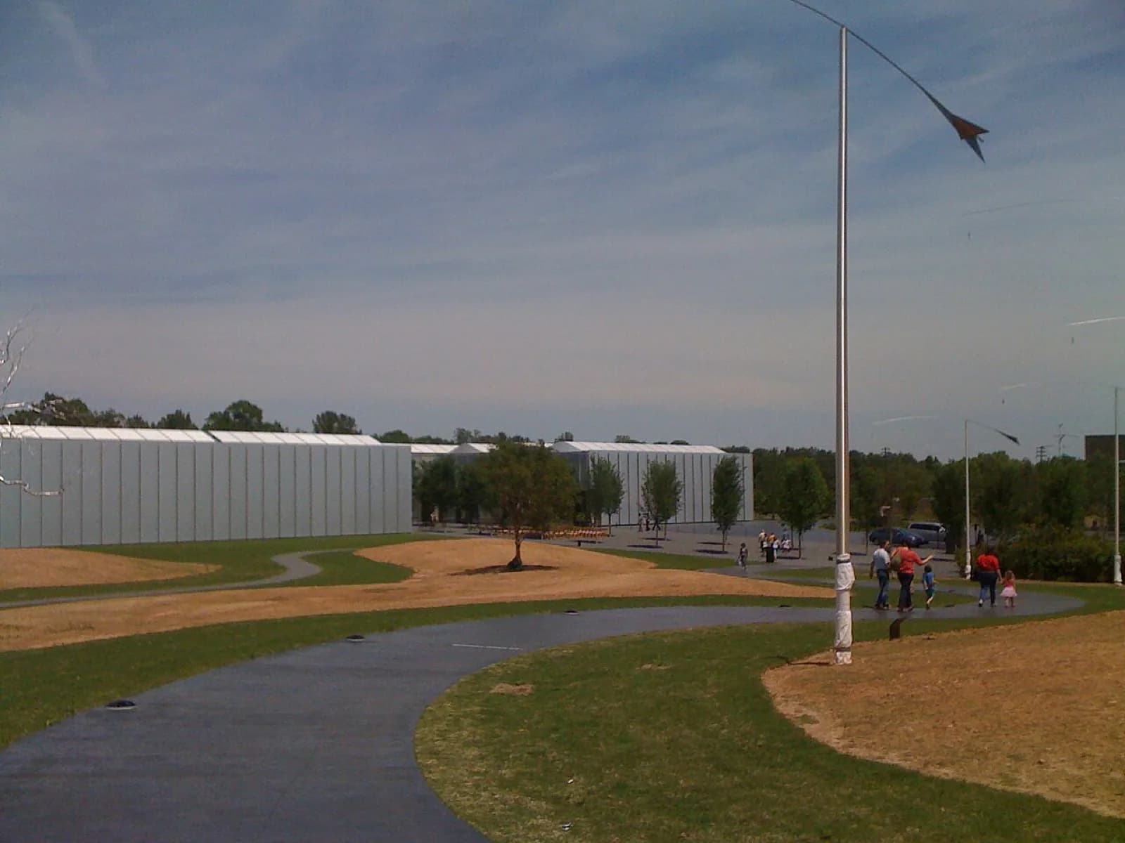 Exterior view of the modern West Building of the North Carolina Museum of Art, a long low structure with gray paneling, in a landscaped park with curving paths, trees, visitors and a tall sculpture pole in the foreground, Raleigh, North Carolina.