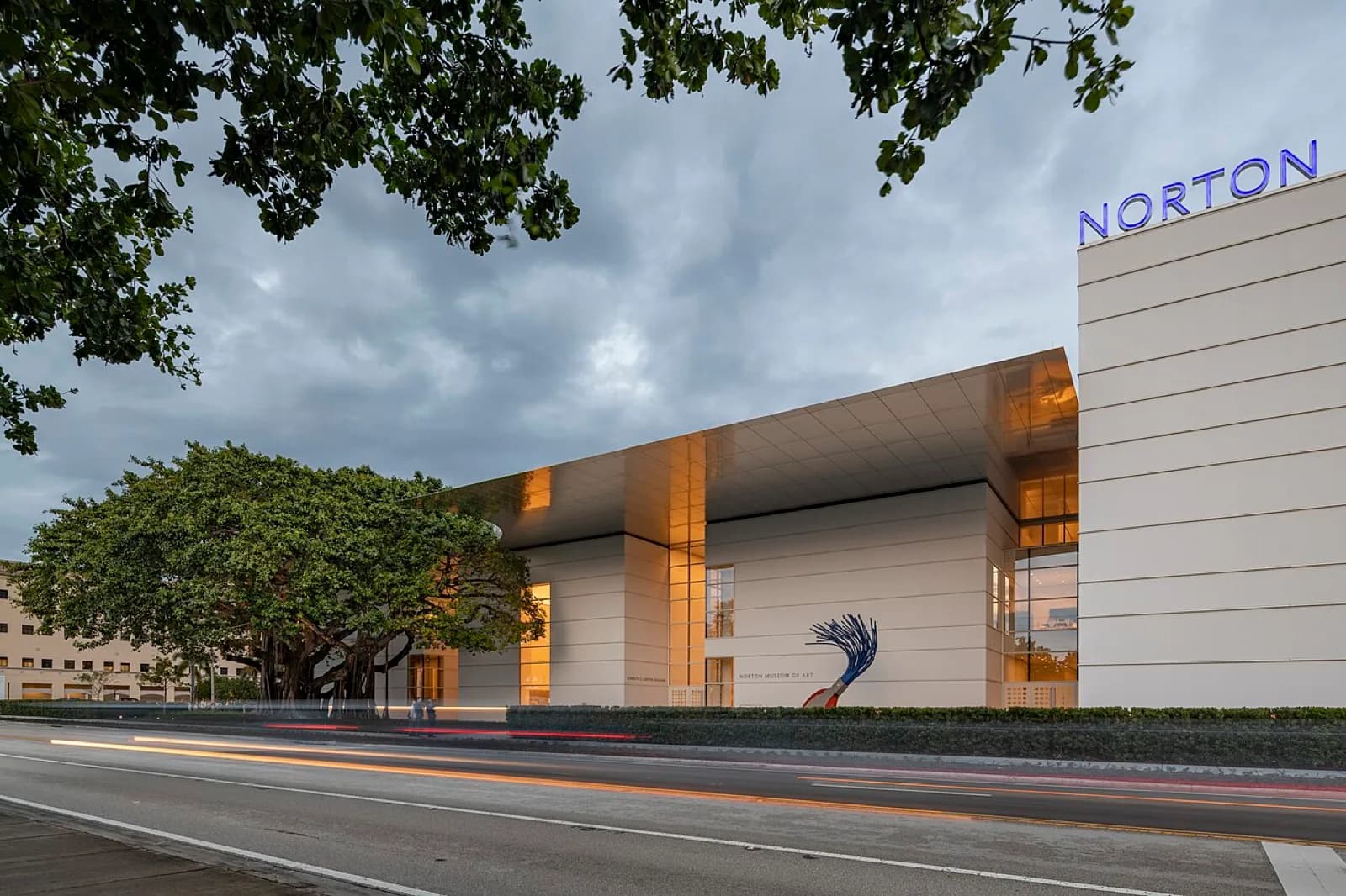 Front facade of the redesigned Norton Museum of Art at dusk, a modern building by Foster & Partners with large glass elements, lit entrance, palm trees, sculpture and road in foreground, West Palm Beach, Florida.