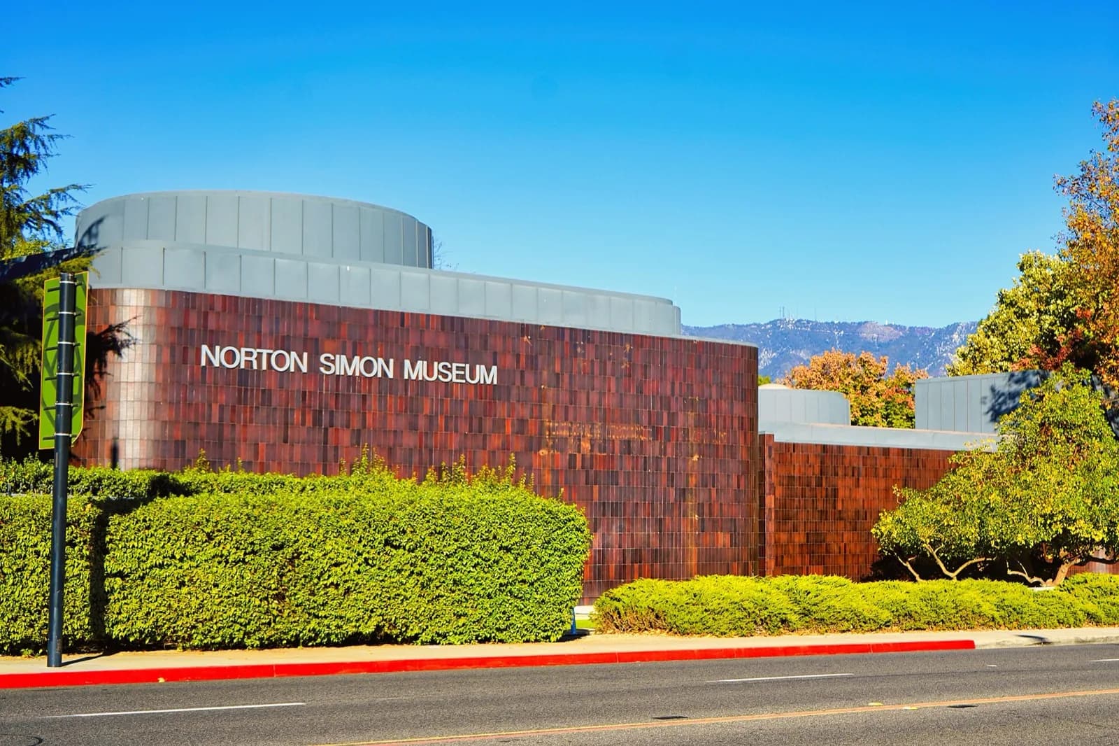 The modern facade of the Norton Simon Museum in Pasadena, featuring its distinctive rusted metal panel walls and curved white architectural elements, surrounded by lush green landscaping with trees and hedges under a clear blue sky with distant mountains.