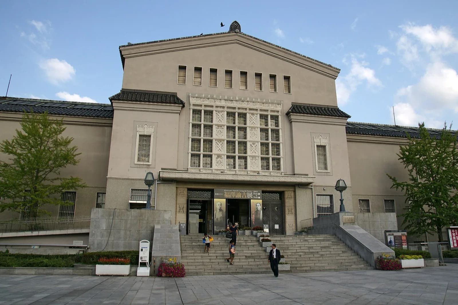 Exterior of the Osaka City Museum of Fine Arts, a large building with traditional Japanese tiled roof and white facade, set in Tennoji Park with trees, lawn and stone walls, Osaka, Japan.