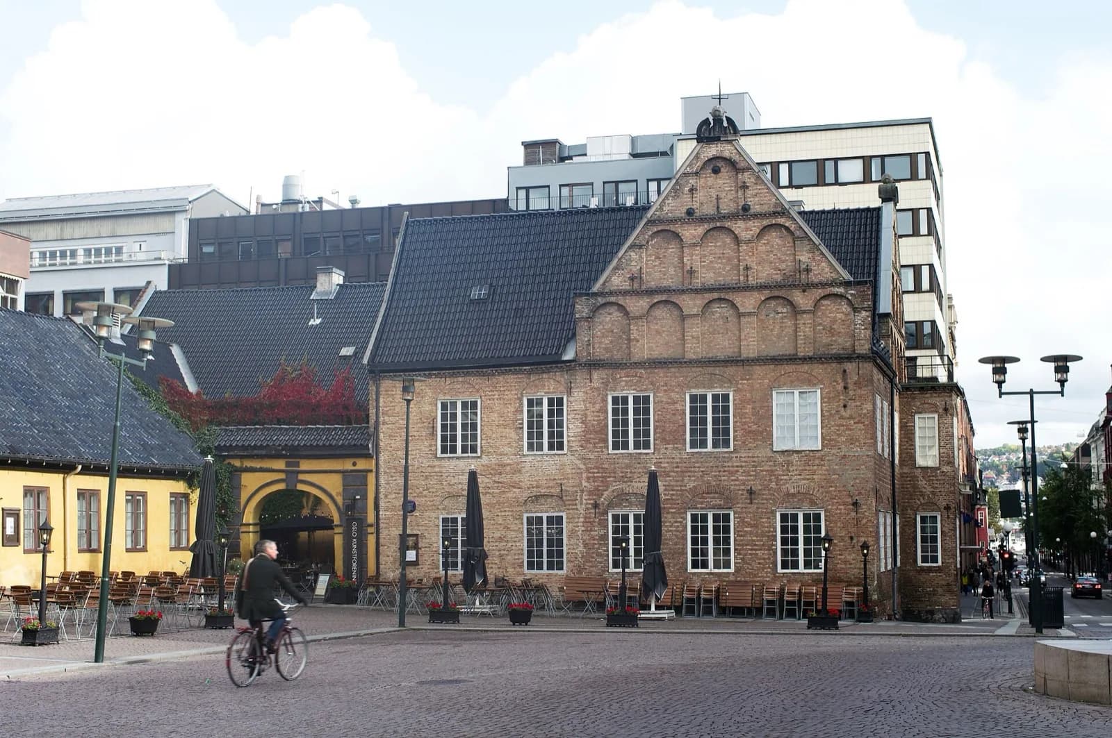 The historic red brick Rådmannsgården building in Oslo, Norway, housing Oslo Kunstforening, with its characteristic stepped gable facade, arched windows, and dark roof, located in a cobblestone square with outdoor seating for a cafe, a cyclist in the foreground, street lamps, and modern buildings behind under a partly cloudy sky.