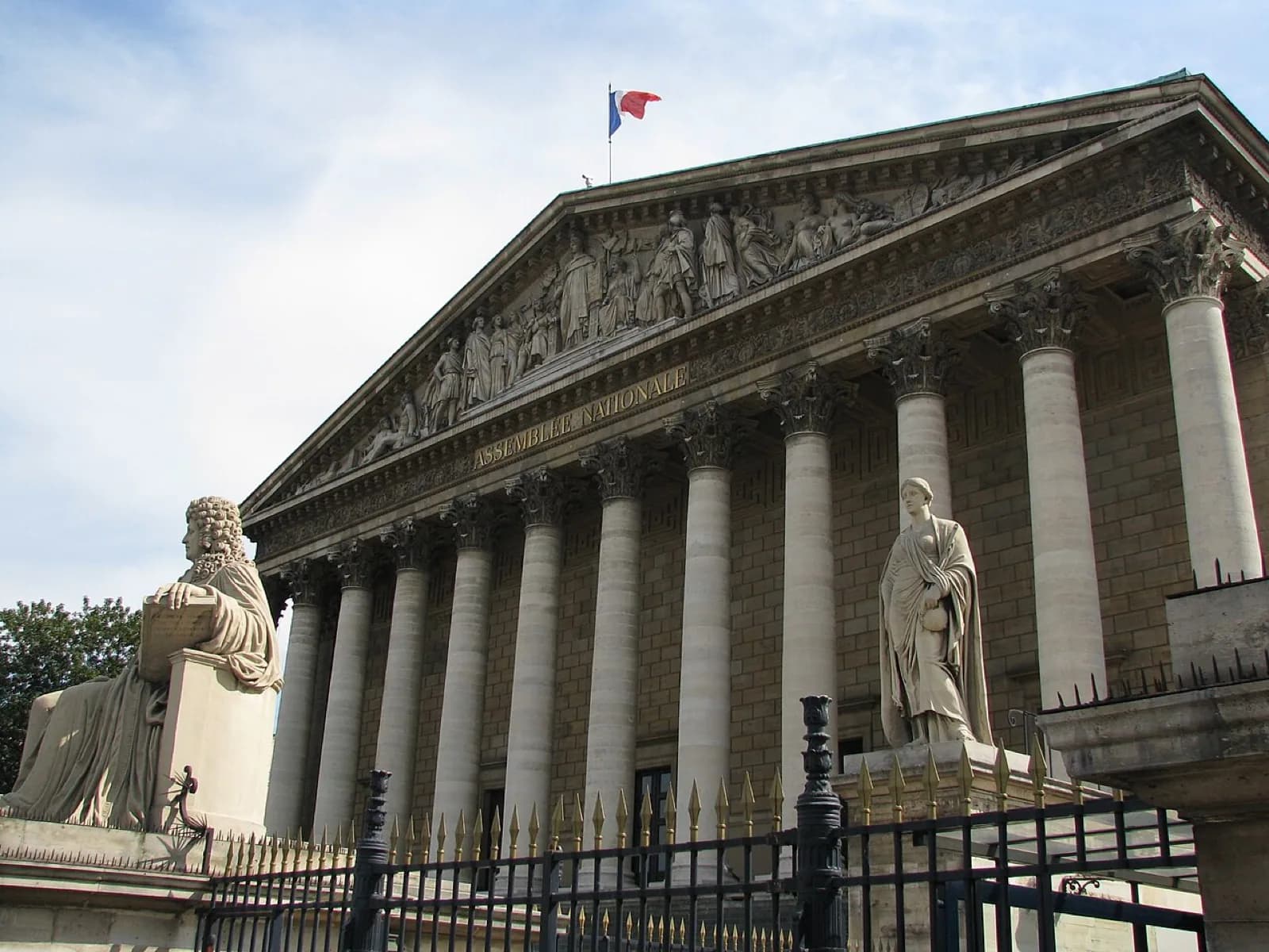 The iconic columned neoclassical facade of the Palais Bourbon, seat of the French National Assembly, with 'ASSEMBLEE NATIONALE' on the pediment, sculptures, French flags, and statues in front, Paris, France.