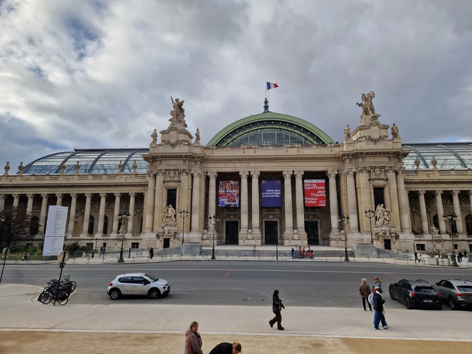 The ornate Beaux-Arts stone facade of the Grand Palais (Palais des Champs-Élysées) in Paris, France, featuring a large central arched entrance under a green glass vaulted roof, classical columns, sculptural pediments, exhibition banners, and a French flag atop, viewed from across the street on an overcast day with pedestrians, vehicles, and bicycles in the foreground.