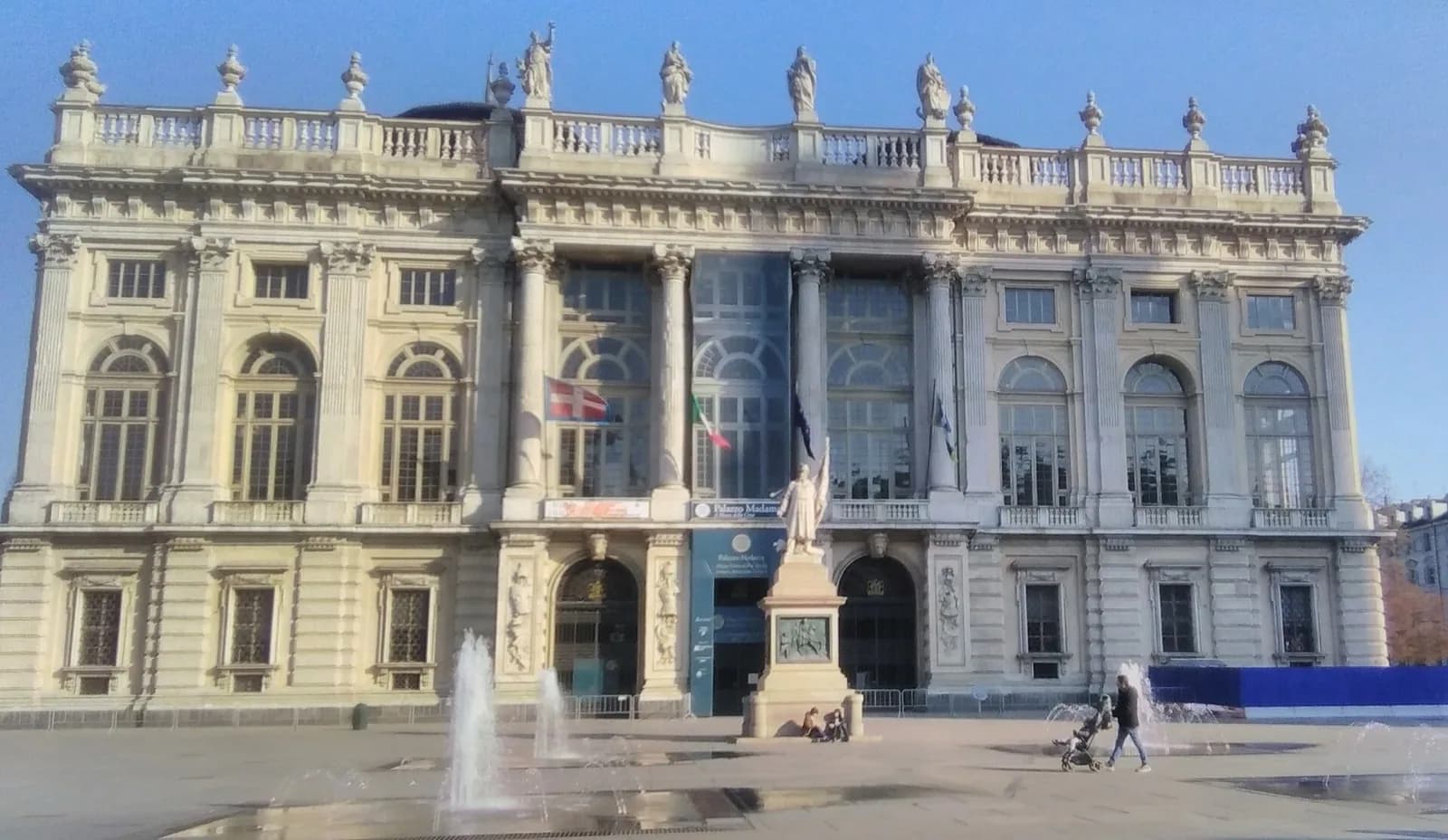 The grand Baroque façade of Palazzo Madama in Turin, Italy, designed by Filippo Juvarra in the early 18th century, featuring a monumental staircase, ornate windows and stonework, fronting Piazza Castello with a monument, surrounding buildings, trees and people under a bright blue sky.