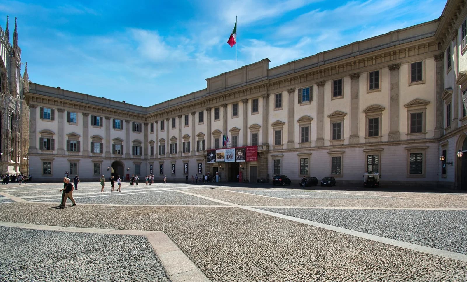 The Palazzo Reale in Milan, a grand neoclassical building with columns and windows, viewed from the cobblestone courtyard with the Italian flag flying on a sunny day.