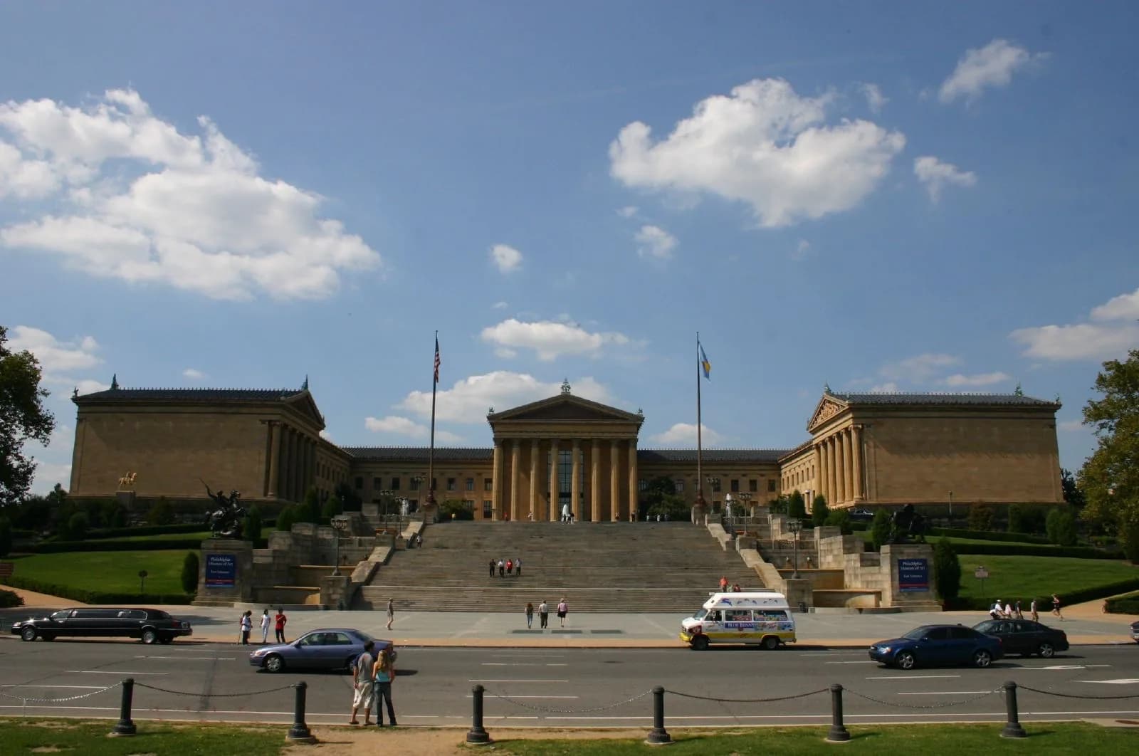 The grand neoclassical facade of the Philadelphia Museum of Art, with its massive columns, pediment, and broad stone steps leading up from the Benjamin Franklin Parkway, under a bright blue sky with clouds.