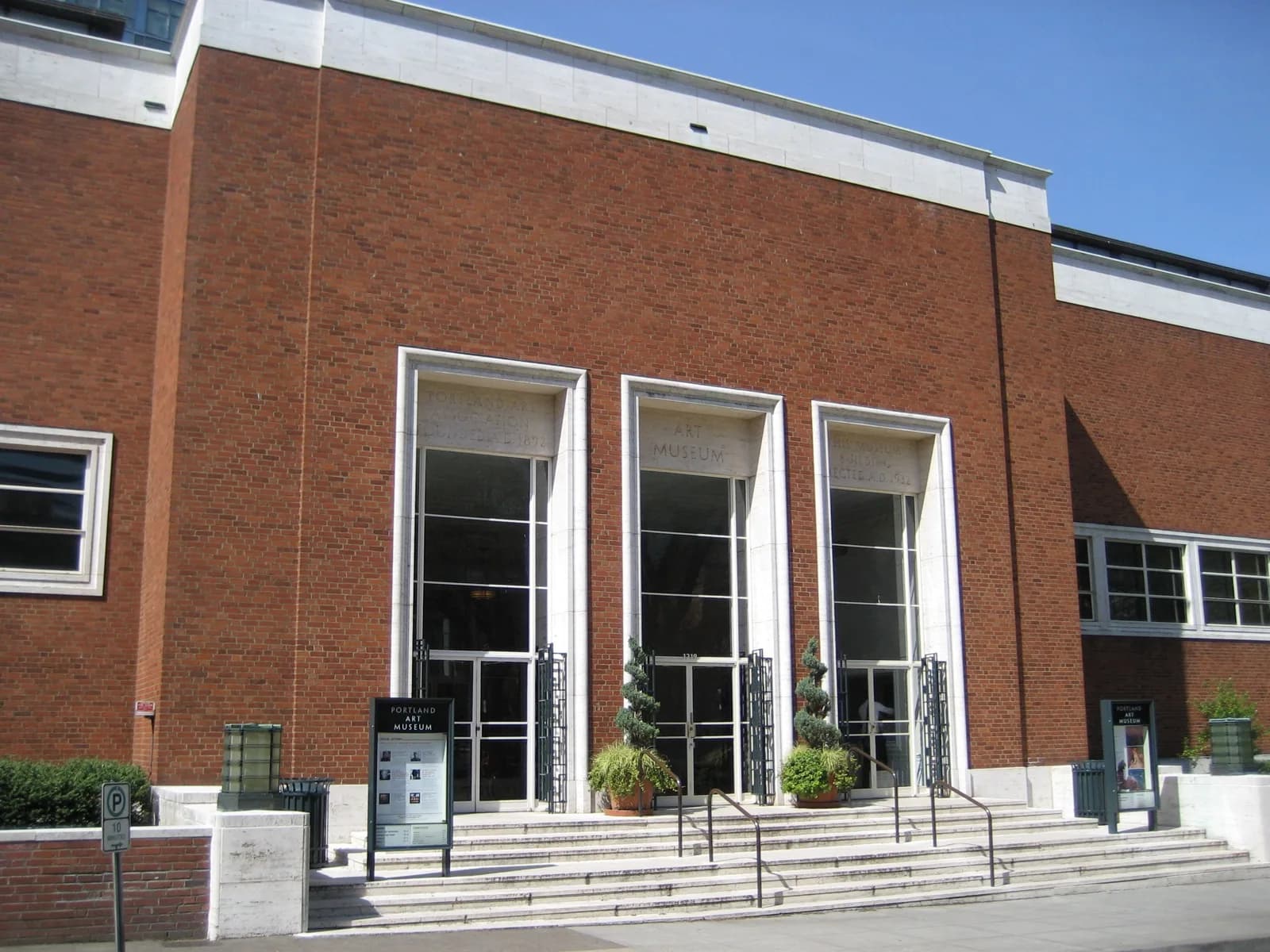 The red brick facade and main entrance of the Portland Art Museum in Portland, Oregon, featuring tall glass doors with carved stone lettering, white trim, and steps leading up under a clear blue sky.