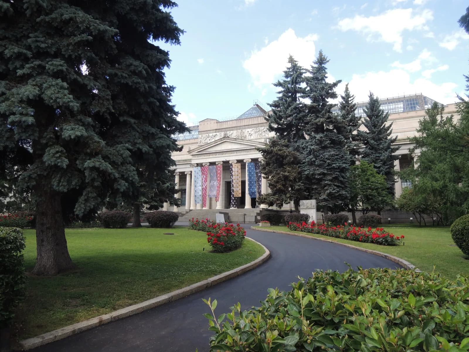 Neoclassical facade of the Pushkin State Museum of Fine Arts in Moscow, featuring a grand portico with tall columns and a sculpted pediment, colorful exhibition banners, surrounded by manicured lawns, mature trees, and red rose beds under a bright blue sky.