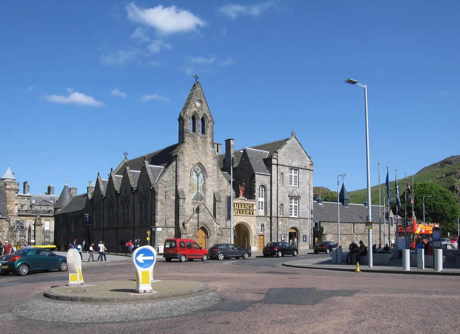 Stone Gothic Revival facade of the Queen's Gallery at the Palace of Holyroodhouse in Edinburgh, featuring a central steeple with cross, arched windows and entrance, prominent 'THE QUEEN'S GALLERY' signage, set against a blue sky with surrounding palace buildings, flags, and urban traffic.
