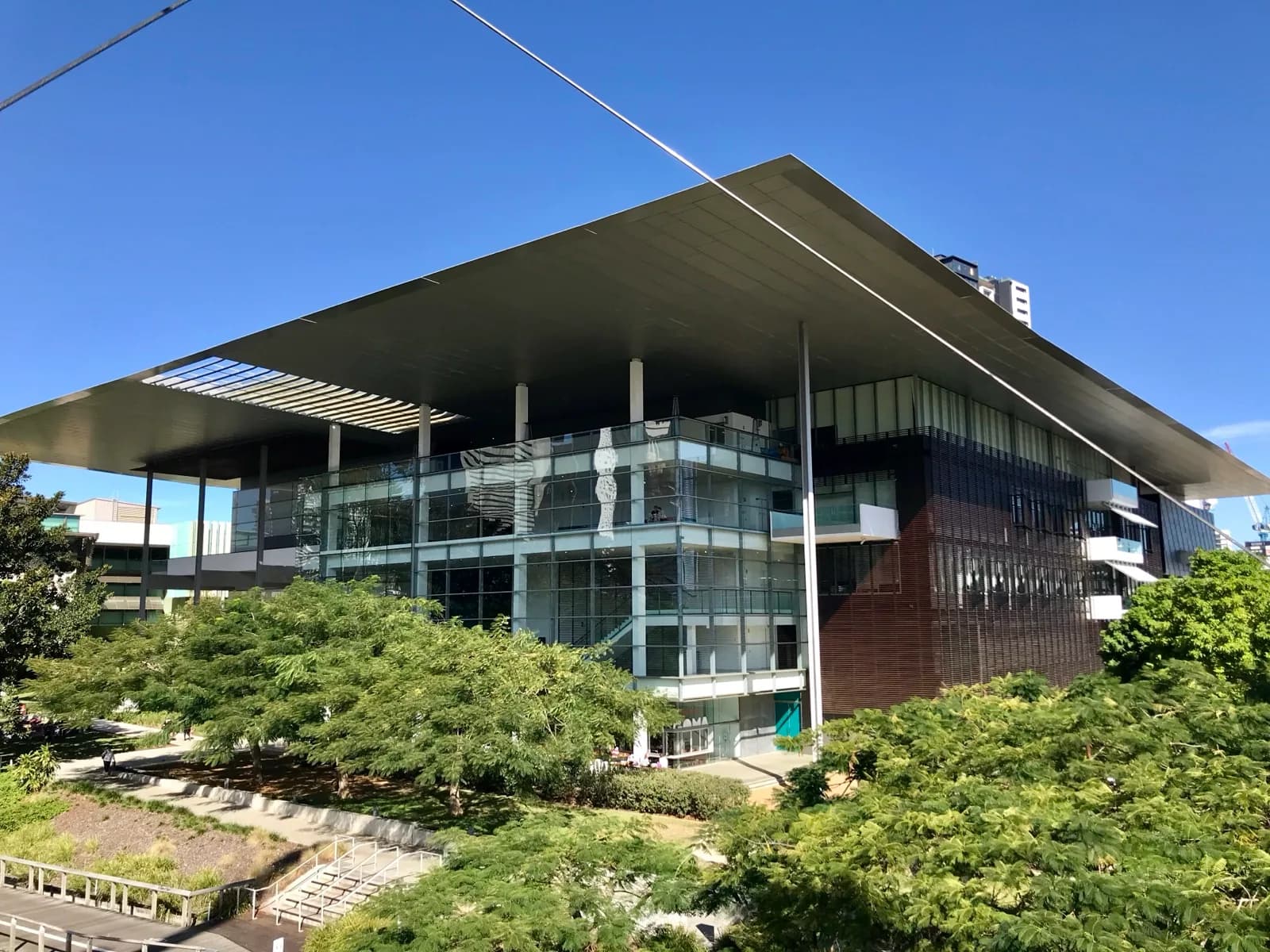 The striking modern facade of the Gallery of Modern Art (GOMA) at QAGOMA in Brisbane, featuring a large cantilevered roof, extensive glass walls, and dark cladding, set amid lush greenery under a clear blue sky, South Bank, Queensland, Australia.