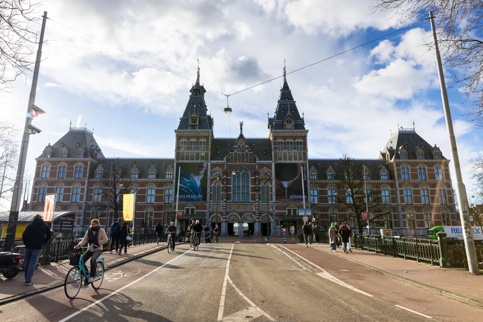 The iconic red-brick Rijksmuseum building in Amsterdam with its twin towers under a blue sky, with Vermeer exhibition banners and visitors in the foreground.